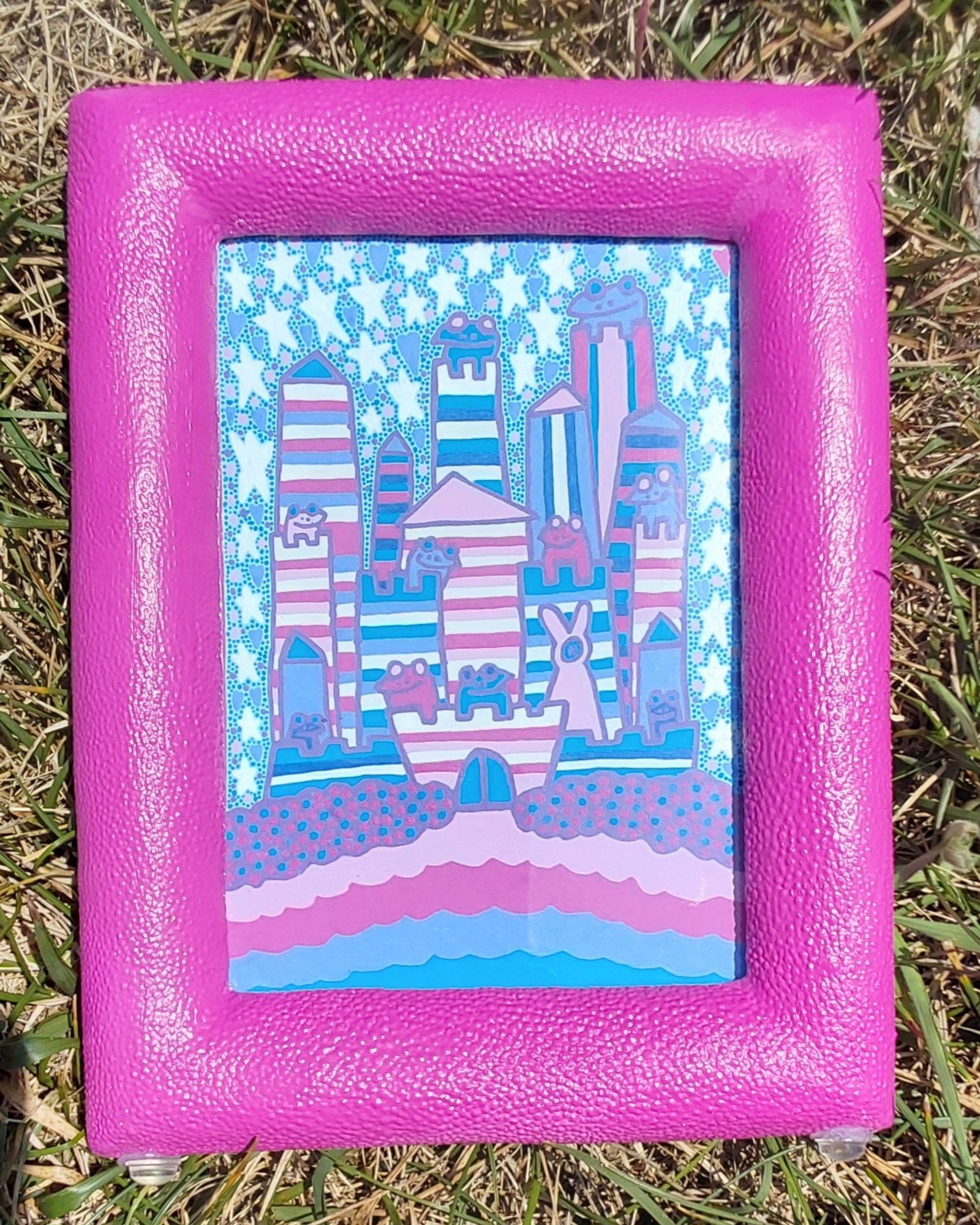 A pink frame surrounds a drawing of a frog castle.
There are 9 frogs and one pink bunny creature. The castle is blue, rose, pink , dark blue, purplish blue, and white. Around the castle, there are white stars, purplish blue hearts, pink, purple, and rose dots. Below the castle are purple bushes with rose and blue berries. 
You can get this piece for 65 shipping included. It is a painted frame 5x7 piece.