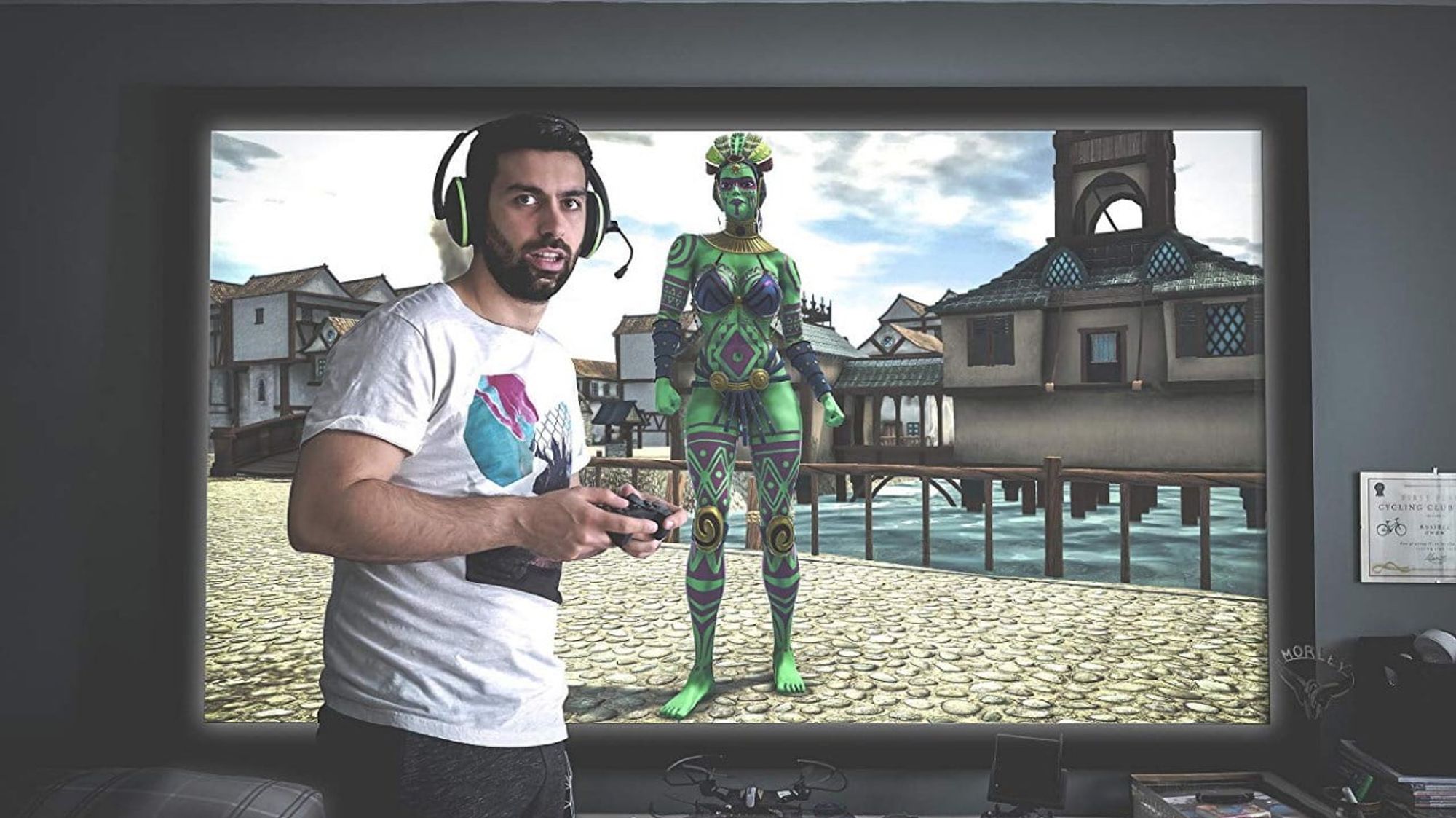 Russell from Dead Pixels standing in front of a screen showing their video game character, a tall green skinned Amazon.