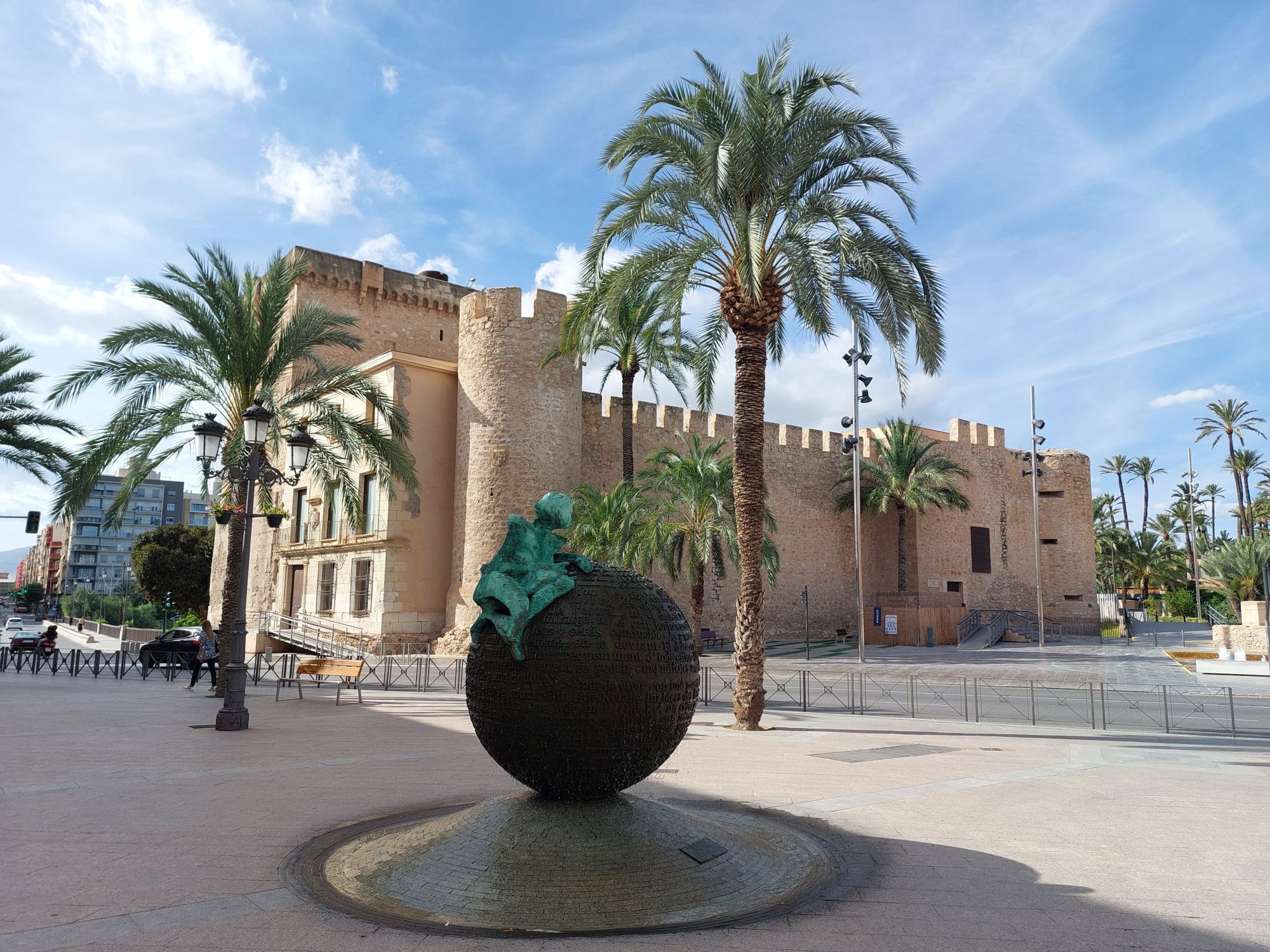 Der Palacio de Altamira unter blauem Himmel und umrahmt von Palmen. Im Vordergrund ein Denkmal.