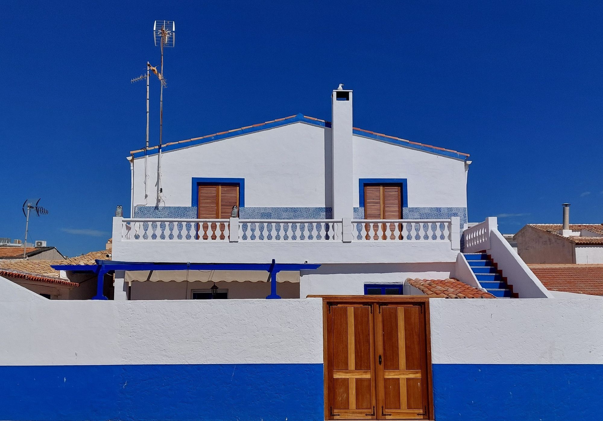 Ein blau-weißes Häuschen mit braunen Fensterläden und Türen, davor eine weiß-blaue Mauer, wobei die blaue Mauerfarbe ziemlich genau der Farbe des wolkenlosen blauen Himmels entspricht.