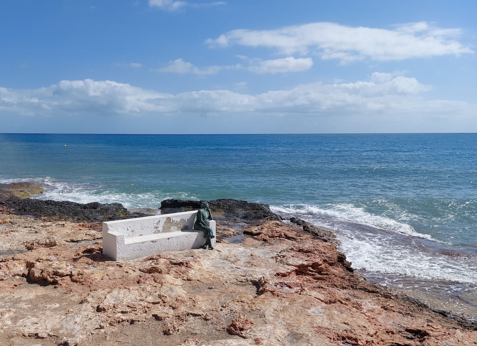 Ein felsiger Mittelmeerstrand. Auf dem Strandfelsen eine weiße Bank aus Stein, auf der als Statue eine nachdenkende Frau sitzt und aufs Meer schaut.