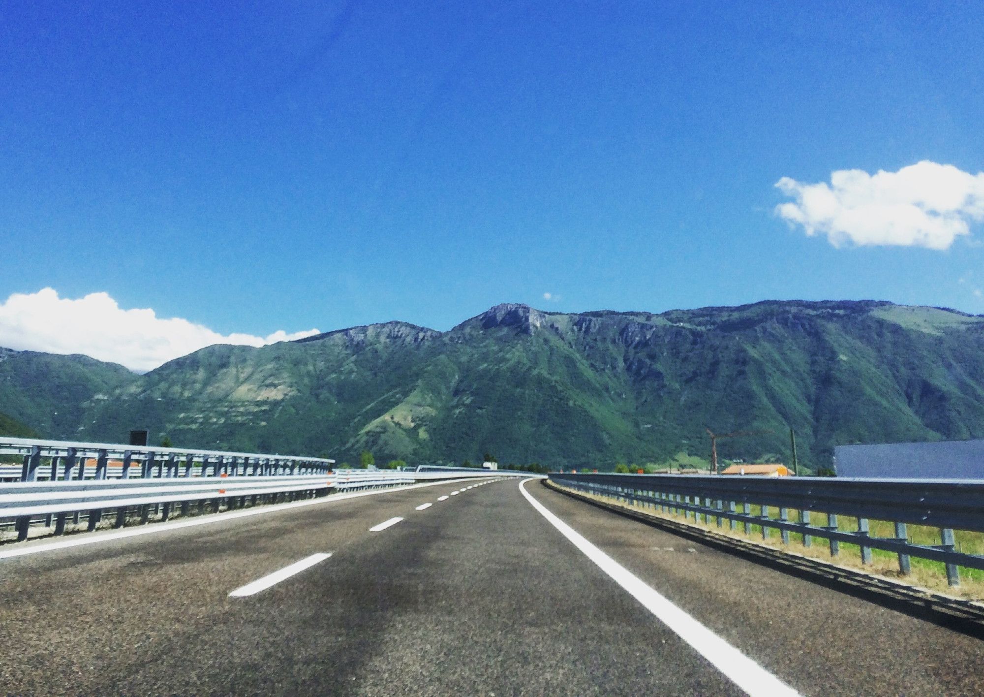 Eine Autobahn in Italien, die auf einen Berg zuführt. Links und rechts Leitplanken. Über dem Berg ein blauer Himmel.