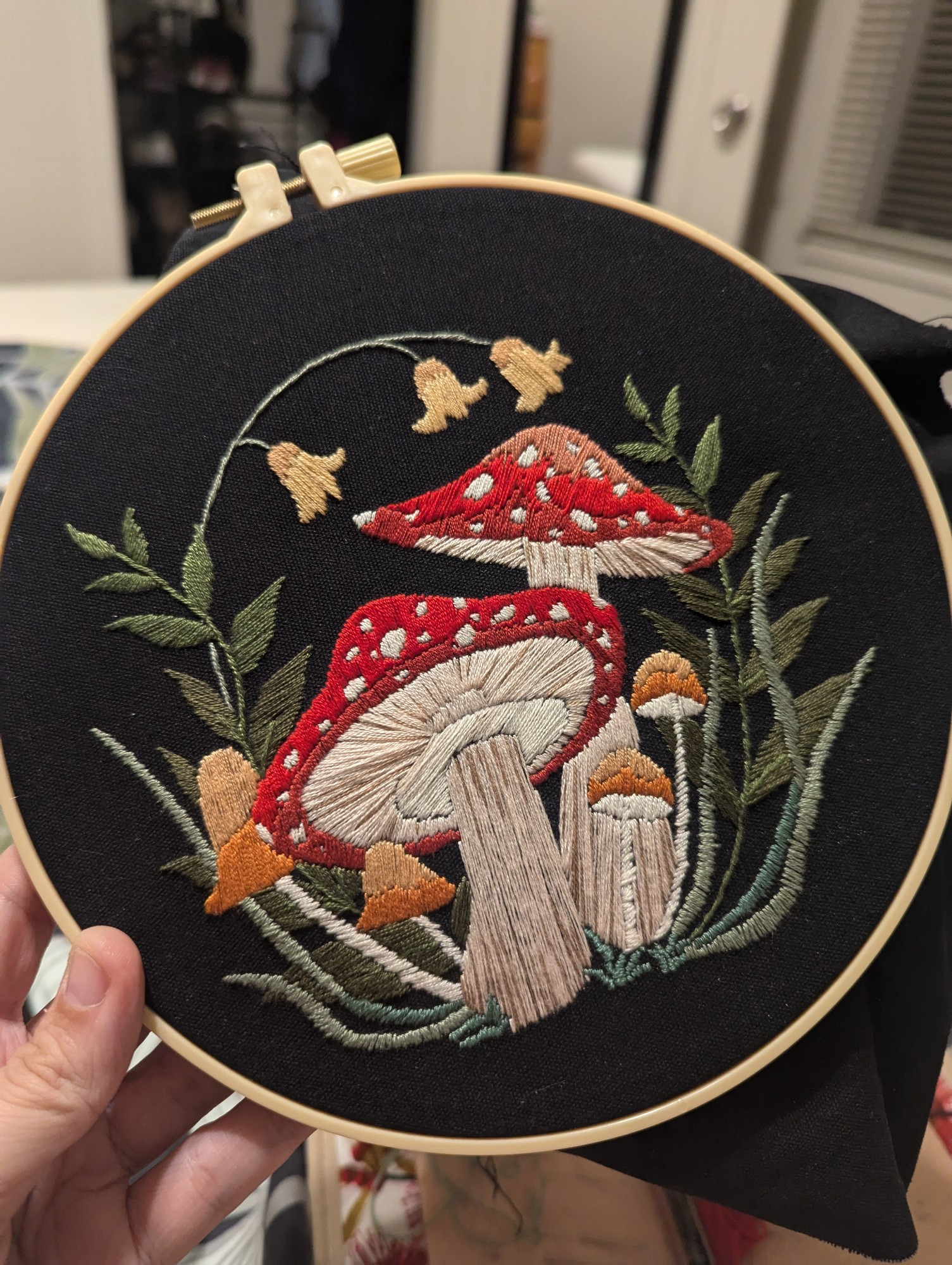 An embroidery hoop with black fabric and an embroidery of two toadstools with small mushrooms and bell-shaped flowers