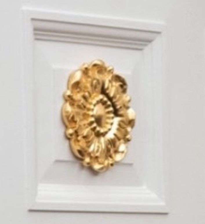 A close up of the one of the gilted doodads Trump's had glued to the doors. It shows that it's several inches larger than the door's raised inset, so the edges extend awkwardly beyond the wood, attached to nothing. 