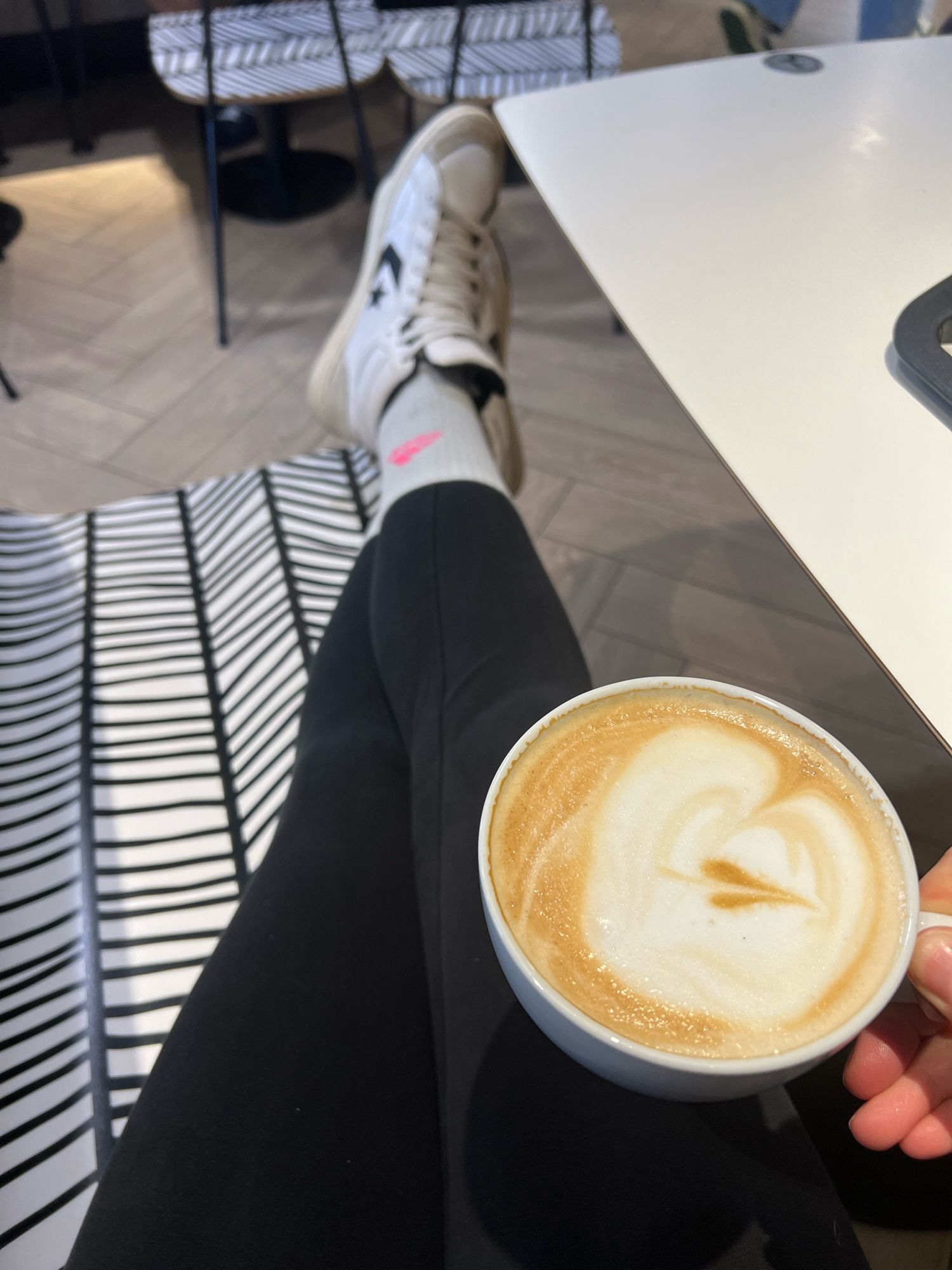 Beine in Schwarzer leggings, tennissocken mit pinkem Logo, Hightopsneaker auf schwarz weiß abstrakter Bank. In der rechten Hand eine große Tasse Capuccino.