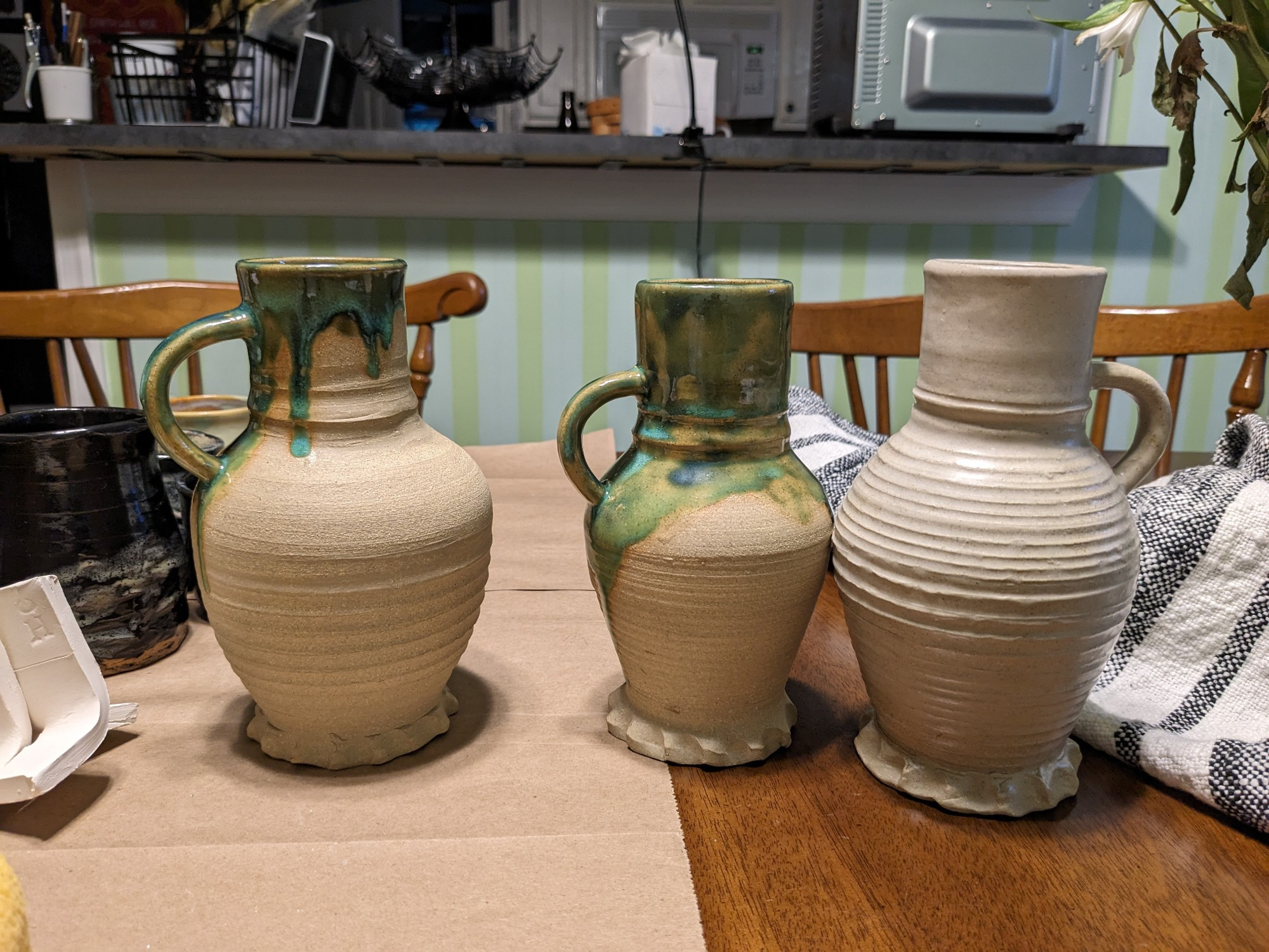 Three buff-coloured jugs with piecrust feet, based on 15th century styles. Two have mottled green and blue glazing on their spouts and handles. The third has clear glaze applied too thick, which lends a milky quality to it. 