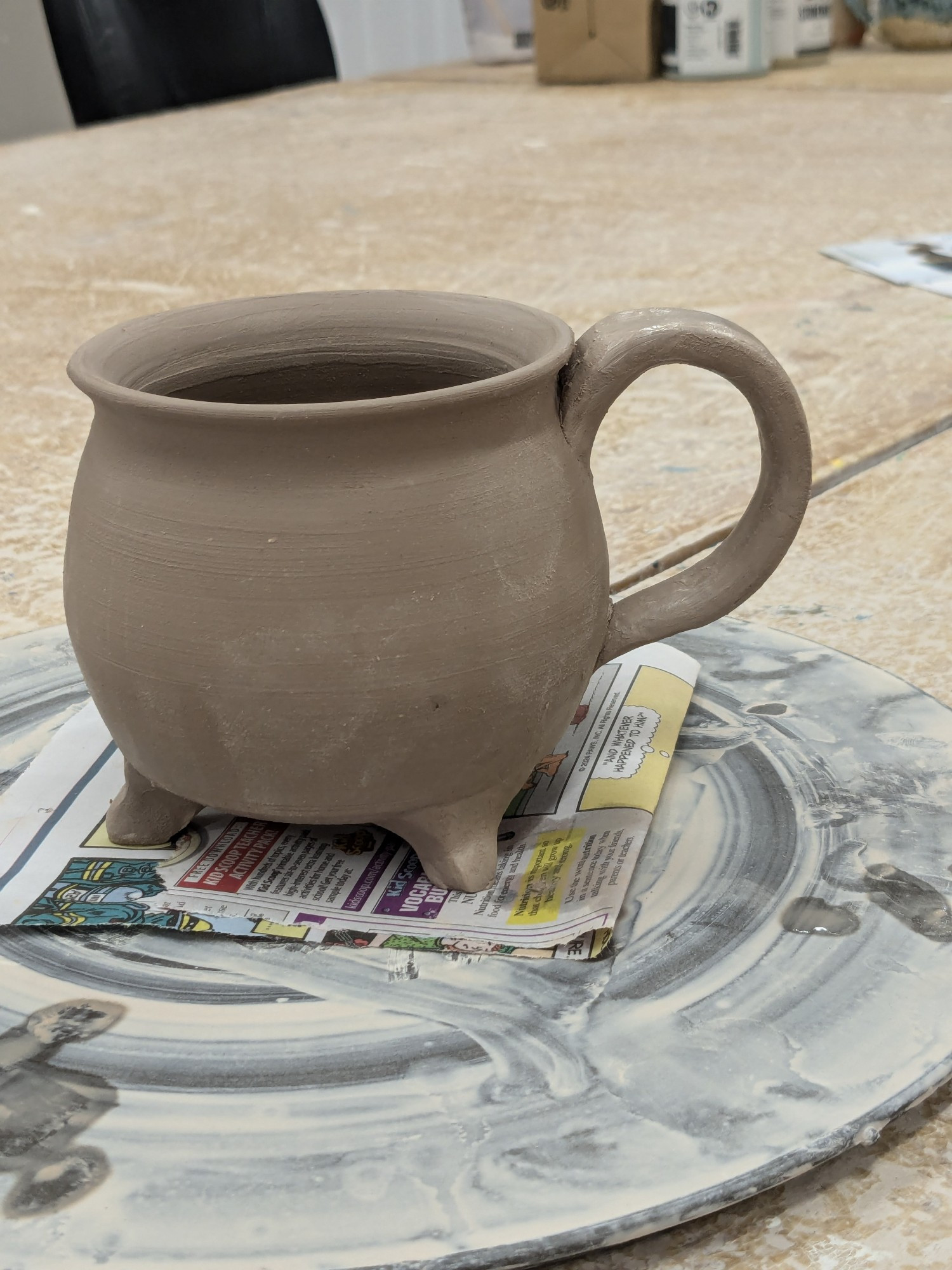 A cauldron-shaped mug on a throwing bat, with a handle coming off the right hand side. The clay is in a leather hard state.