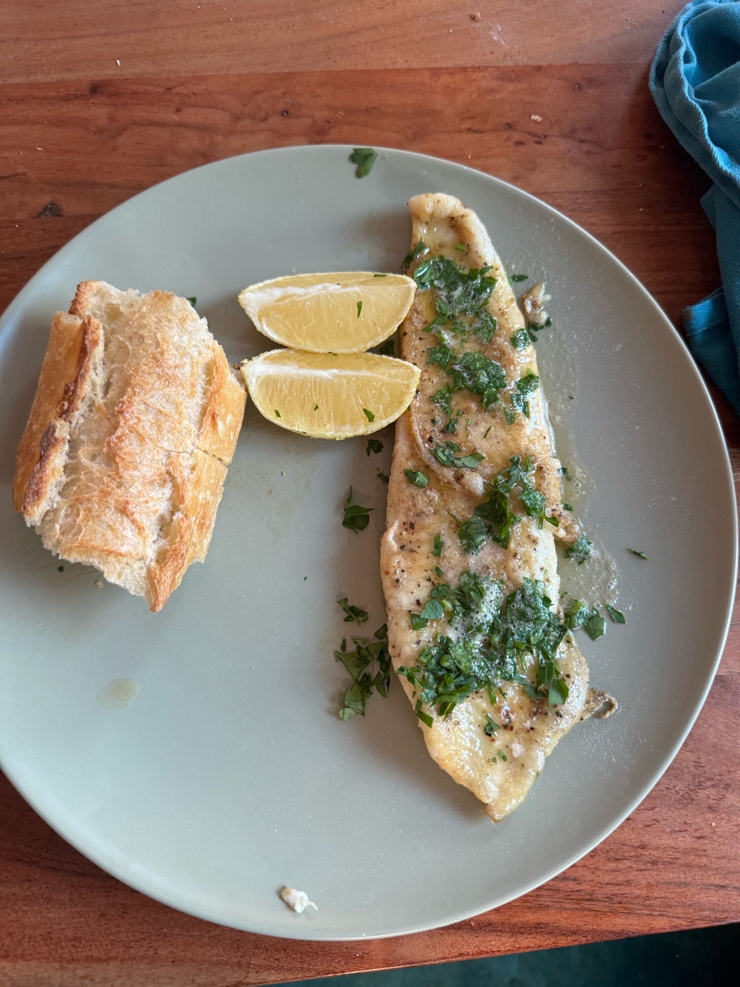 A golden brown fish fillet on a plate, covered in parsley. There’s bread and lemon wedges next to it.