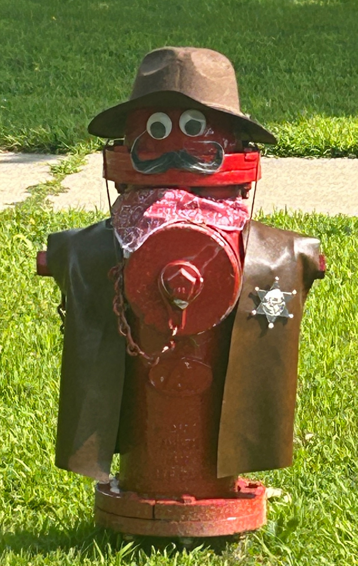 A fire hydrant is dressed up with googly eyes, a faux leather Brown vest with a sheriffs badge on it, and a ranger-type hat