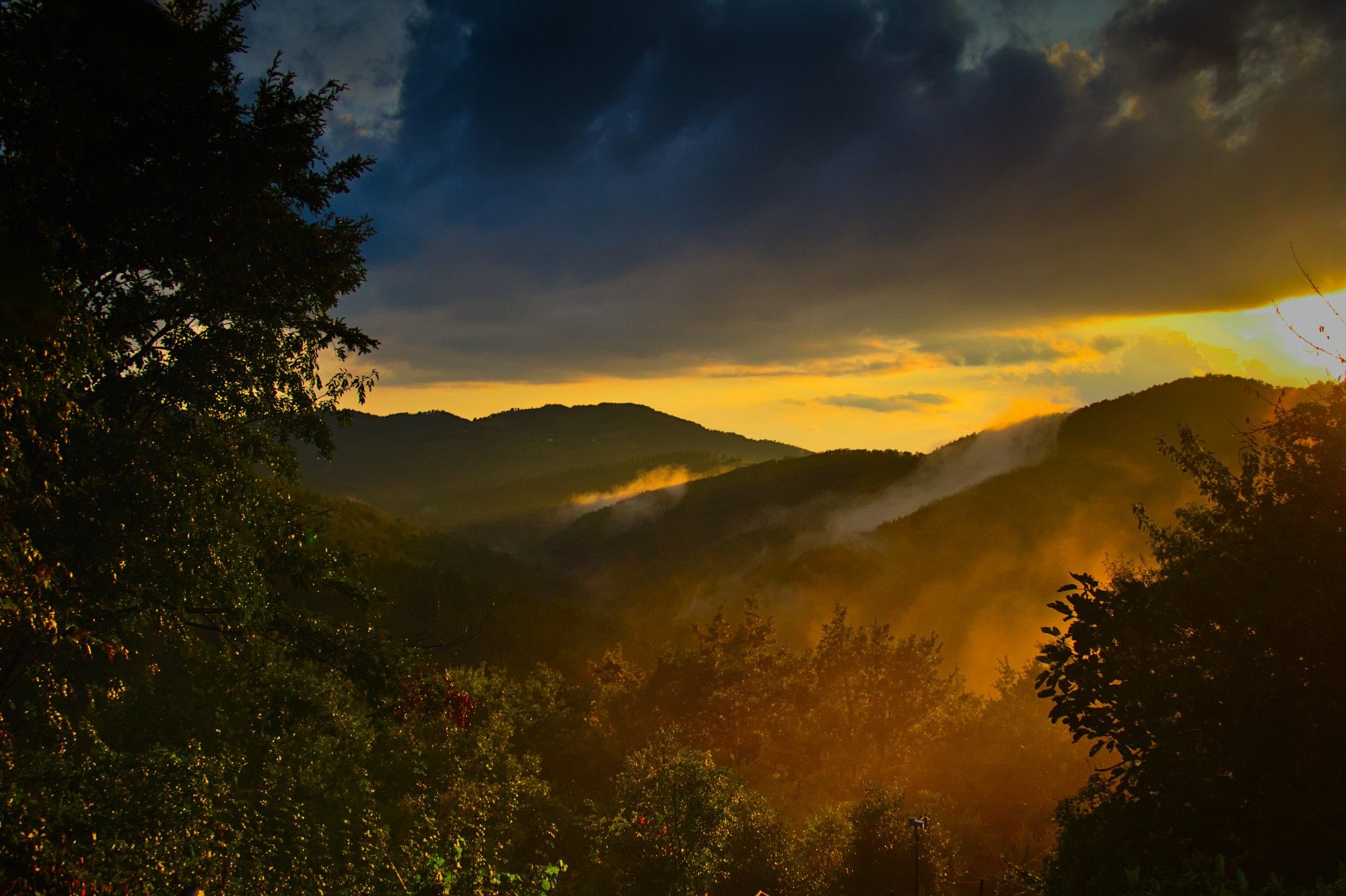 a dark misty valley illuminated in orange light by the setting sun on the right of the frame under a dark cloudy sky