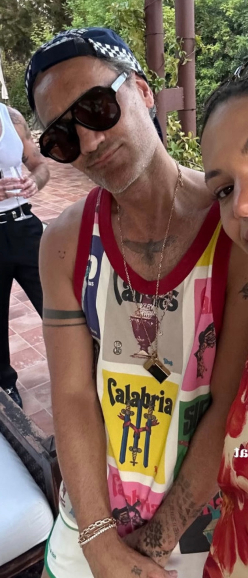 Taika standing with his arms in front of him with his head tilted smiling. He’s wearing a police cap and tank top with red banding around the arms and neck. It’s got a vintage poster collage of various items, one is a sundae and another 3 swords (I think). He has a long chain necklace with what looks like a cigarette lighter on the end and he’s wearing several silver bracelets on his right wrist. 