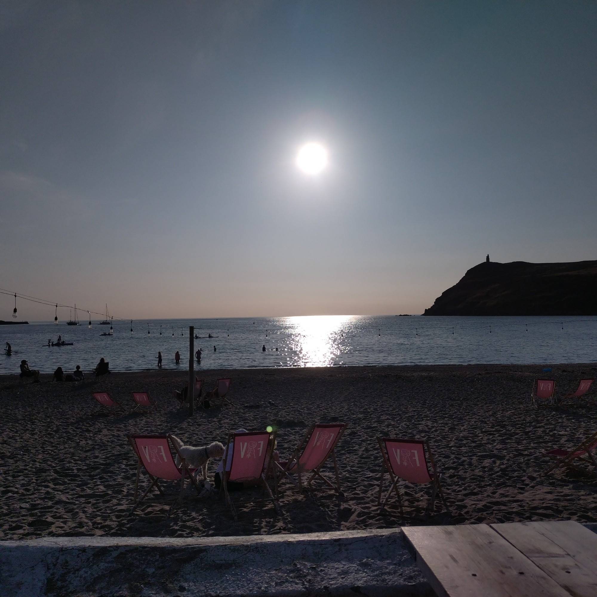 The sun shining above Port Erin beach and Bradda Head . There's some reddish pink deck chairs on the beach in the foreground and the sea reflecting the sunlight below the sky in the background.