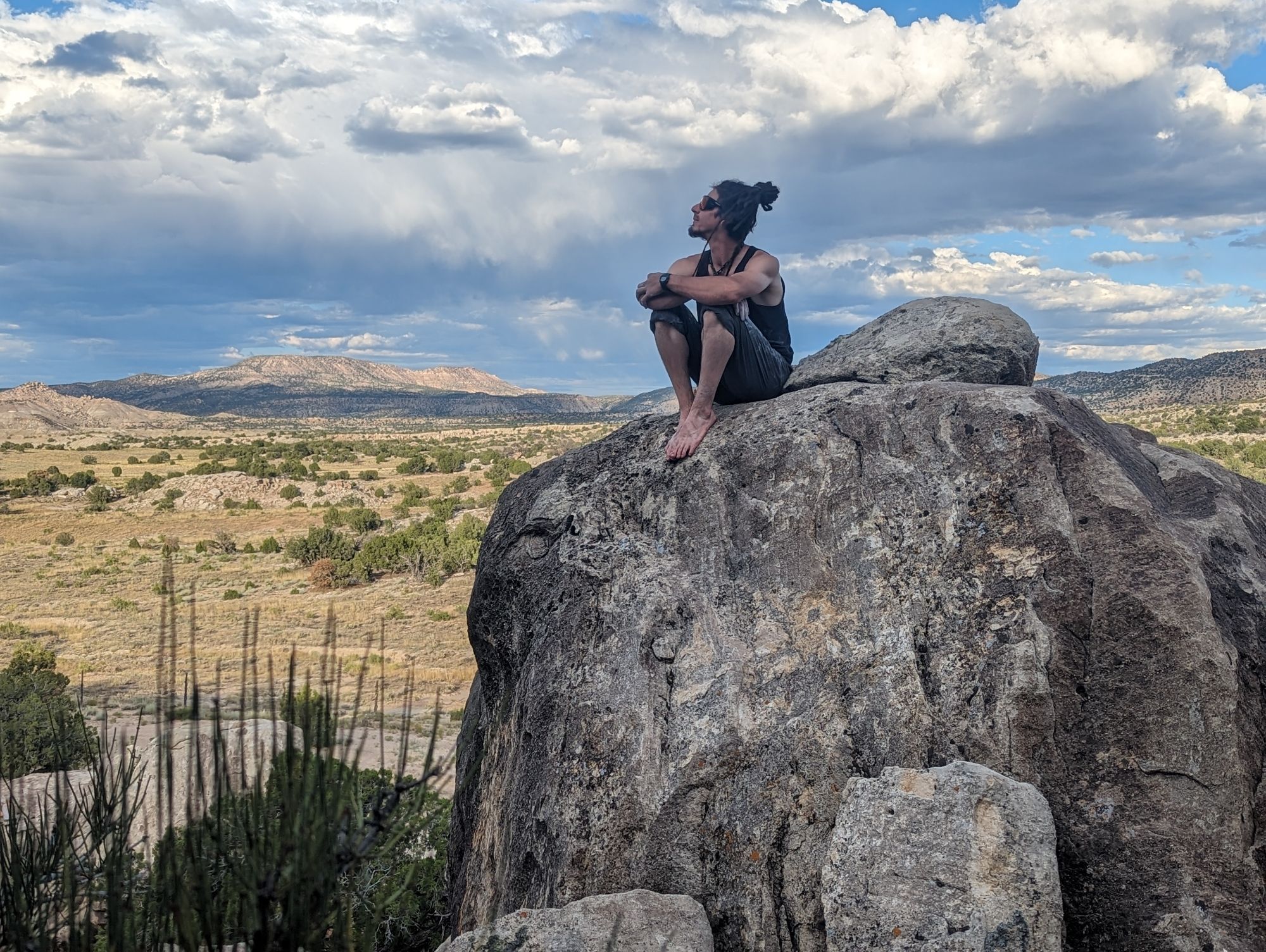 A picture of a guy sitting on a rock looking to his right at the sky. In the background there is a desert landscape and cloud cover