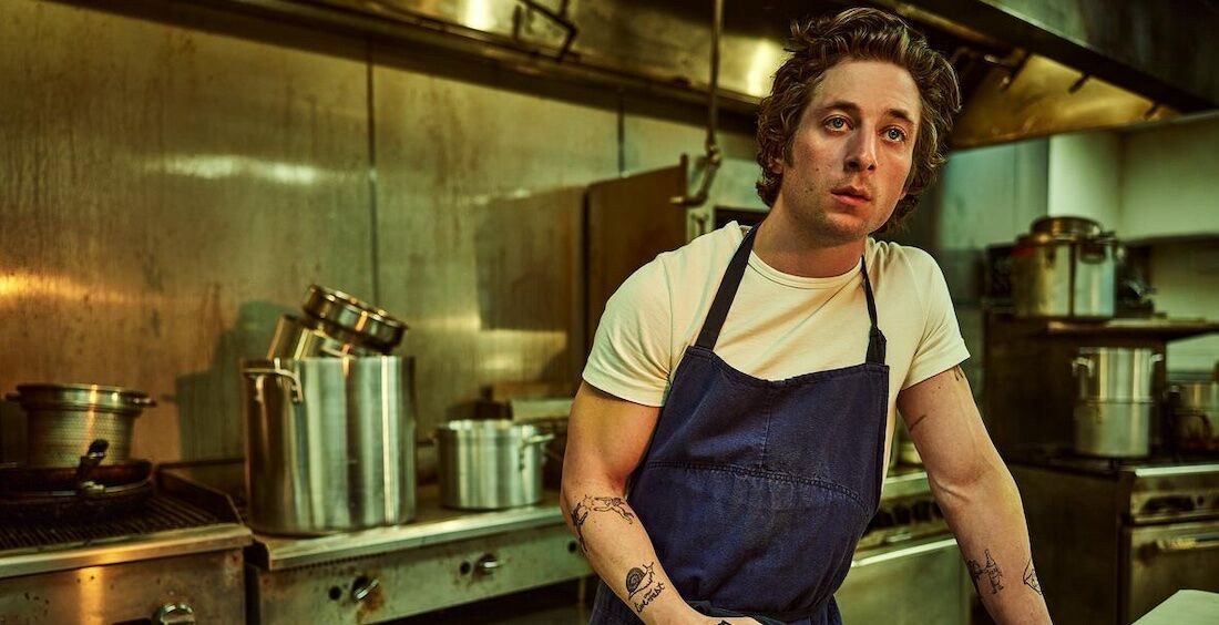 Actor Jeremy Allen White as chef Carmen “Carmy” Berzatto. Here he is standing in the kitchen wearing a white shirt and black apron. Lots of pots stacked up on the stove behind him.