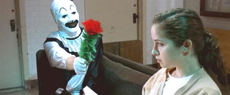 Art the Clown (actor Mike Giannelli) holding a flower towards a woman (actor Kayla Lian)