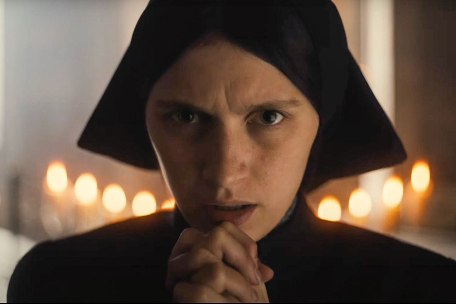 Actor Nell Tiger Free as Margaret. Here she is dressed in all black like a nun, one who hasn’t taken their vows. She is praying with her hands clasped near her chin. Candles lit in the background.