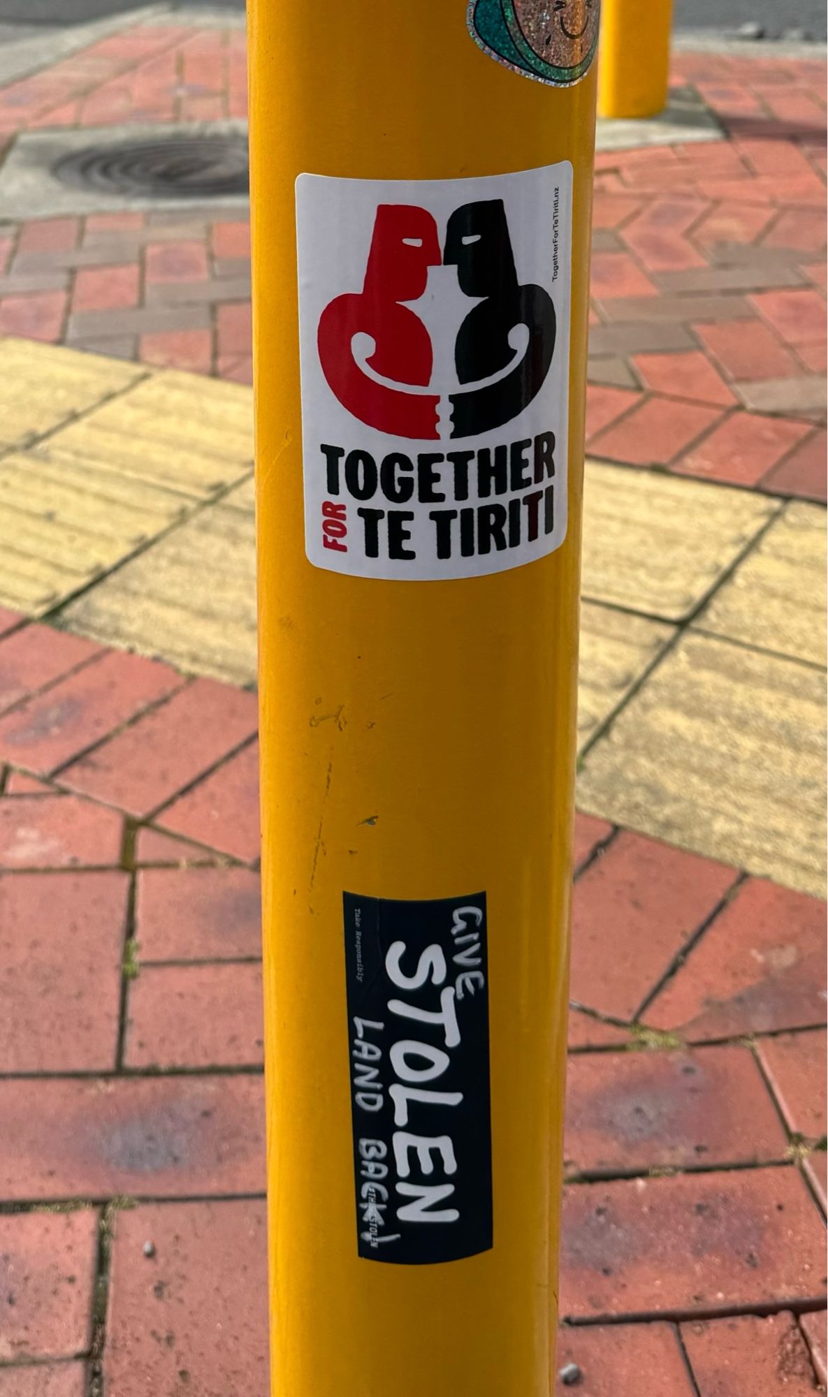 A yellow traffic pole adorned with two stickers: 
Together for Te Tiriti; and
Give Stolen Land Back