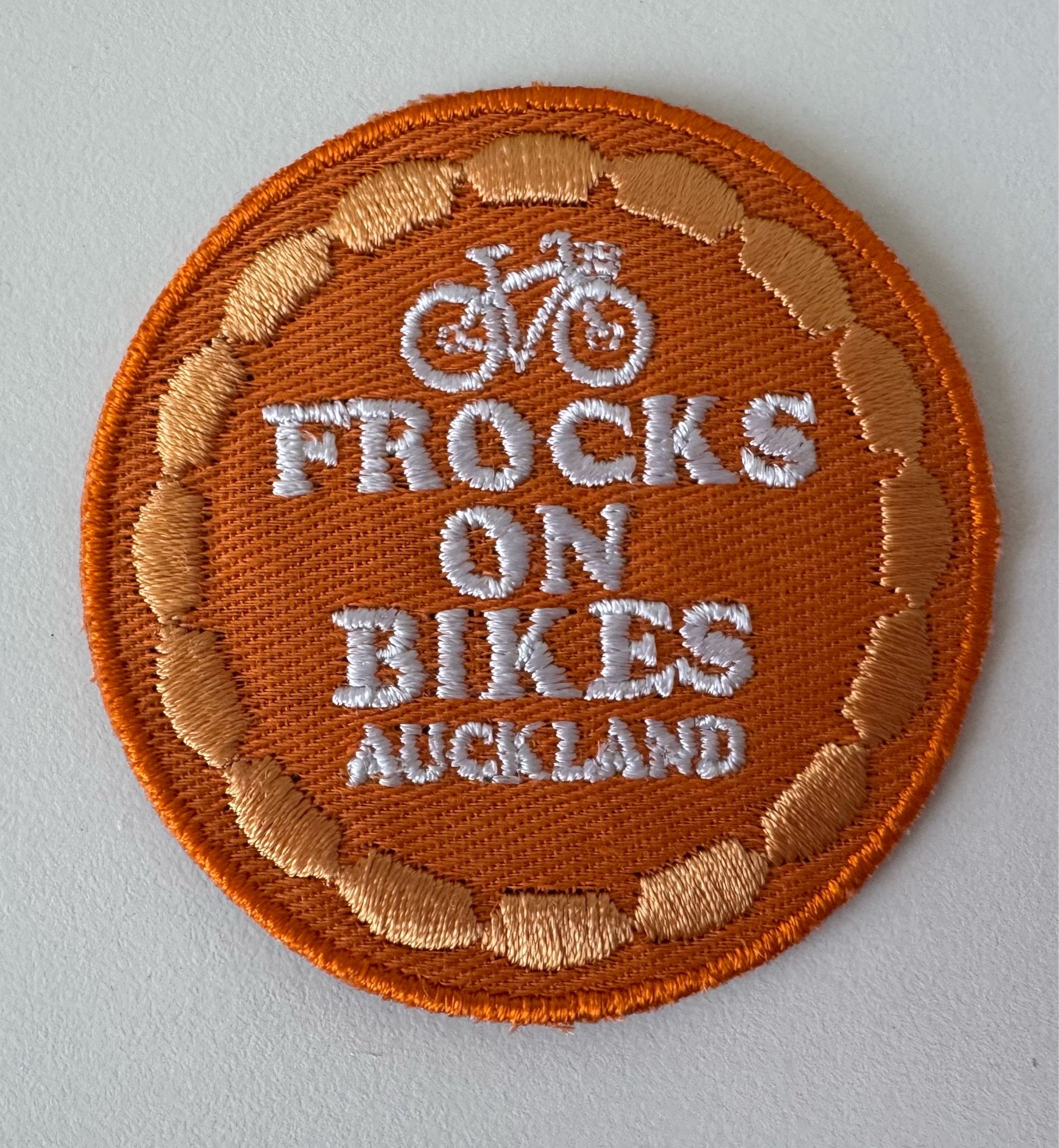 A circle embroidery patch in orange, with a bike and white stitching that reads: Frocks on Bikes Auckland