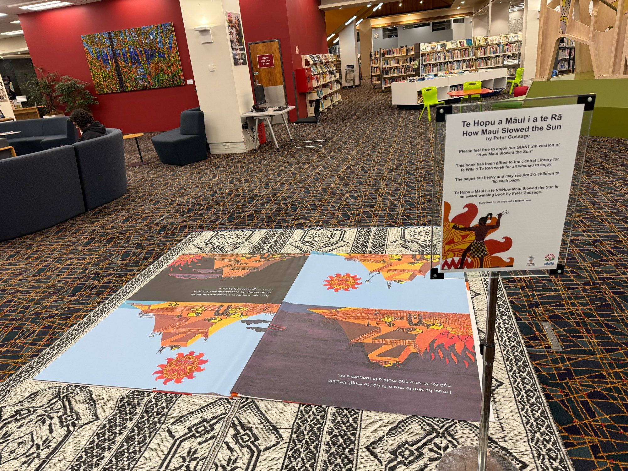 A 2 meter tall cloth book of “Te Hopu a Māui i a te Rā / How Maui Slowed the Sun by Peter Gossage” on a mat in the floor of the Auckland Central Library