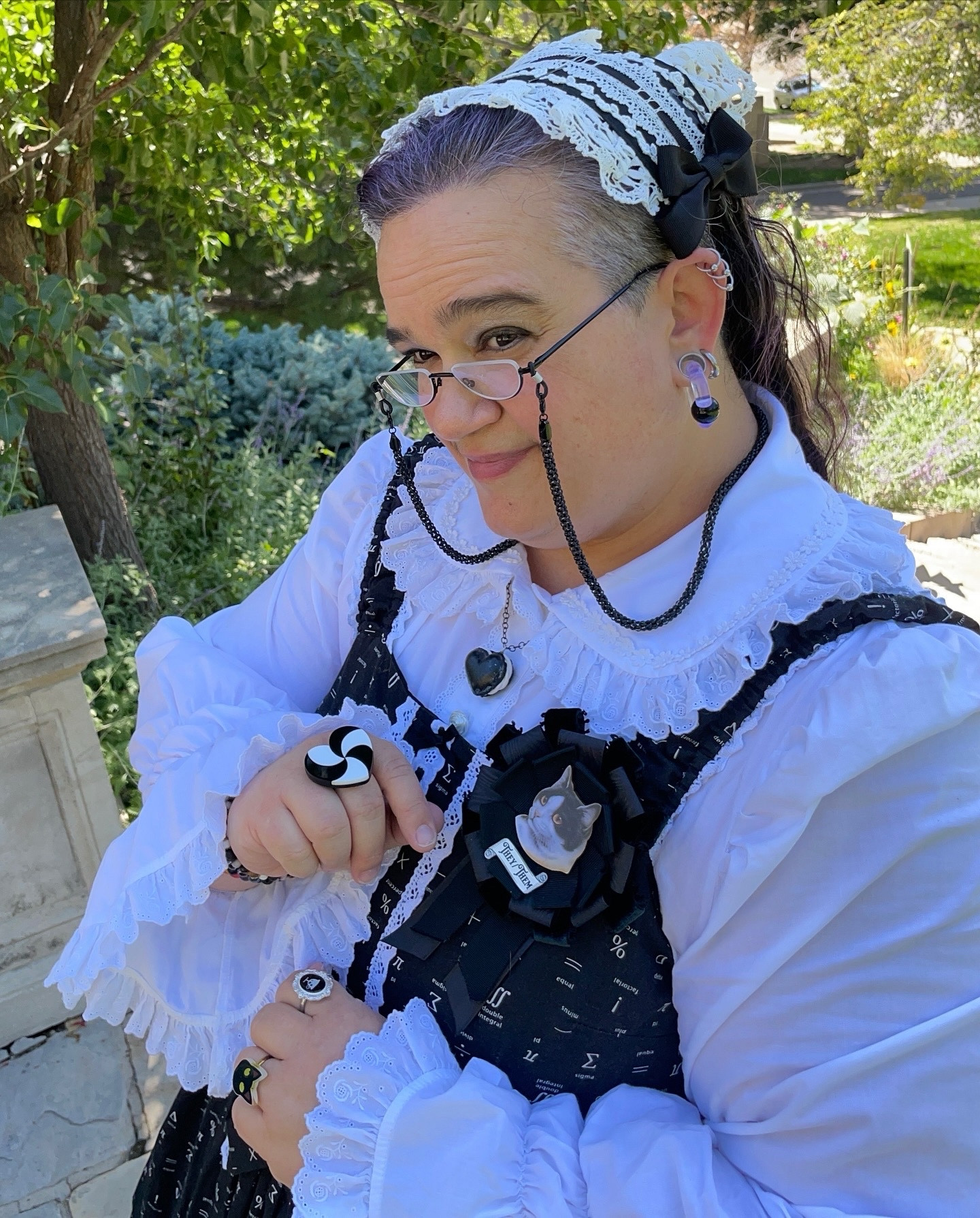 A close up of Elya's outfit, showing details: with the lacy white collar over the math print black and white JSK bodice, they are wearing a heart-shaped macaron pendant, a ribbon rosette with a cat portrait and a scroll reading "they/them", a heart-shaped ring with a swirl pattern, a ring with a tiny painting of a teacup, a ring shaped like a cat's face, half-moon reading glasses with a beaded chain, chonky hoop earrings through tunnels in their lobes, and a lacy headdress; all in the black and white color scheme of the whole outfit.