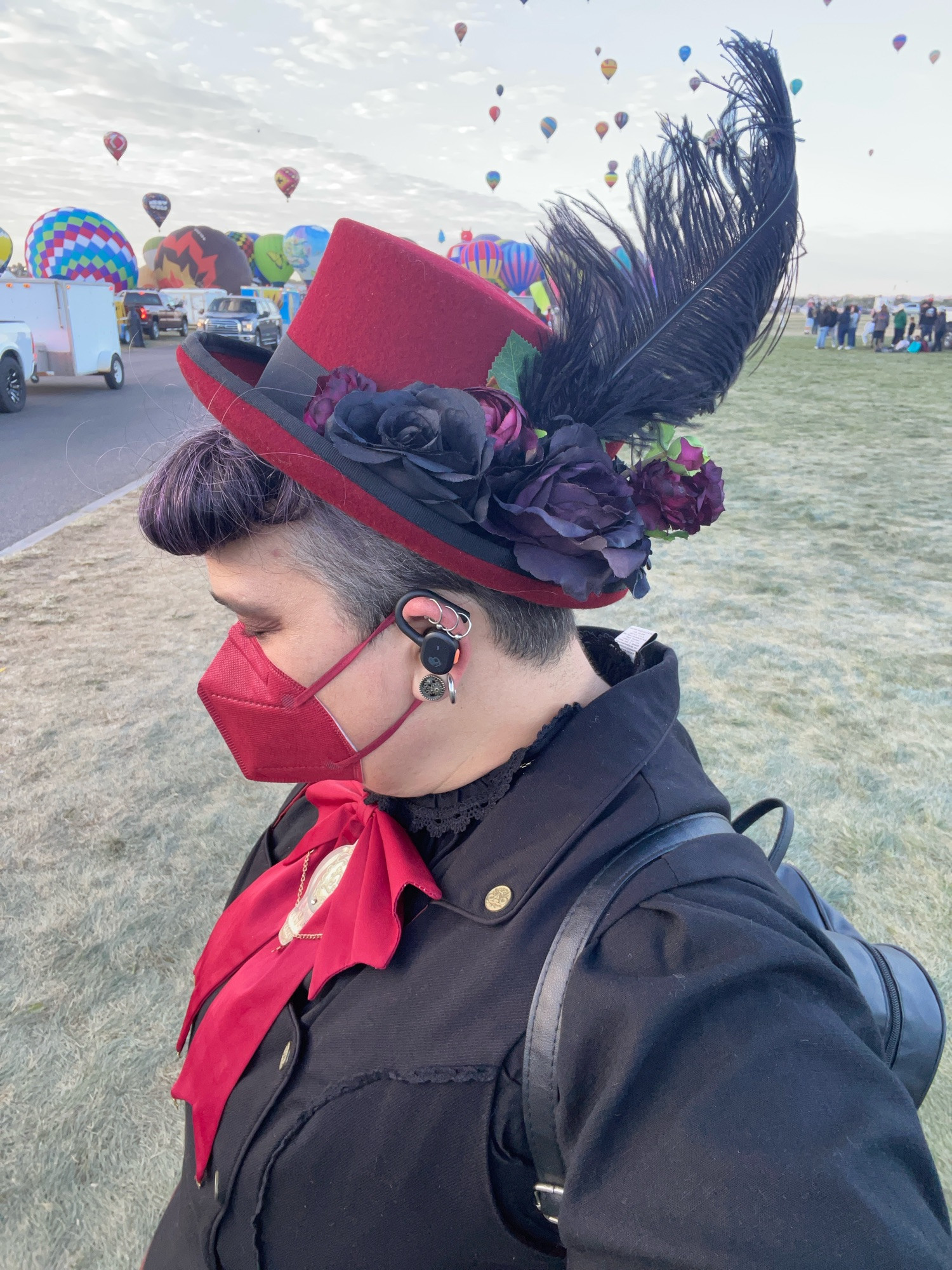 A selfie of Elya with their head turned to the side to show off their magnificent hat, which is a dark red felt short top hat accented with black and dark red roses and a large fluffy black feather. Their hair is styled in bumper bangs. Along with the neck bow, they are wearing a gold-toned balloon shaped pendant. They are also wearing a red KN95 mask color-coordinated with the rest of their outfit. 