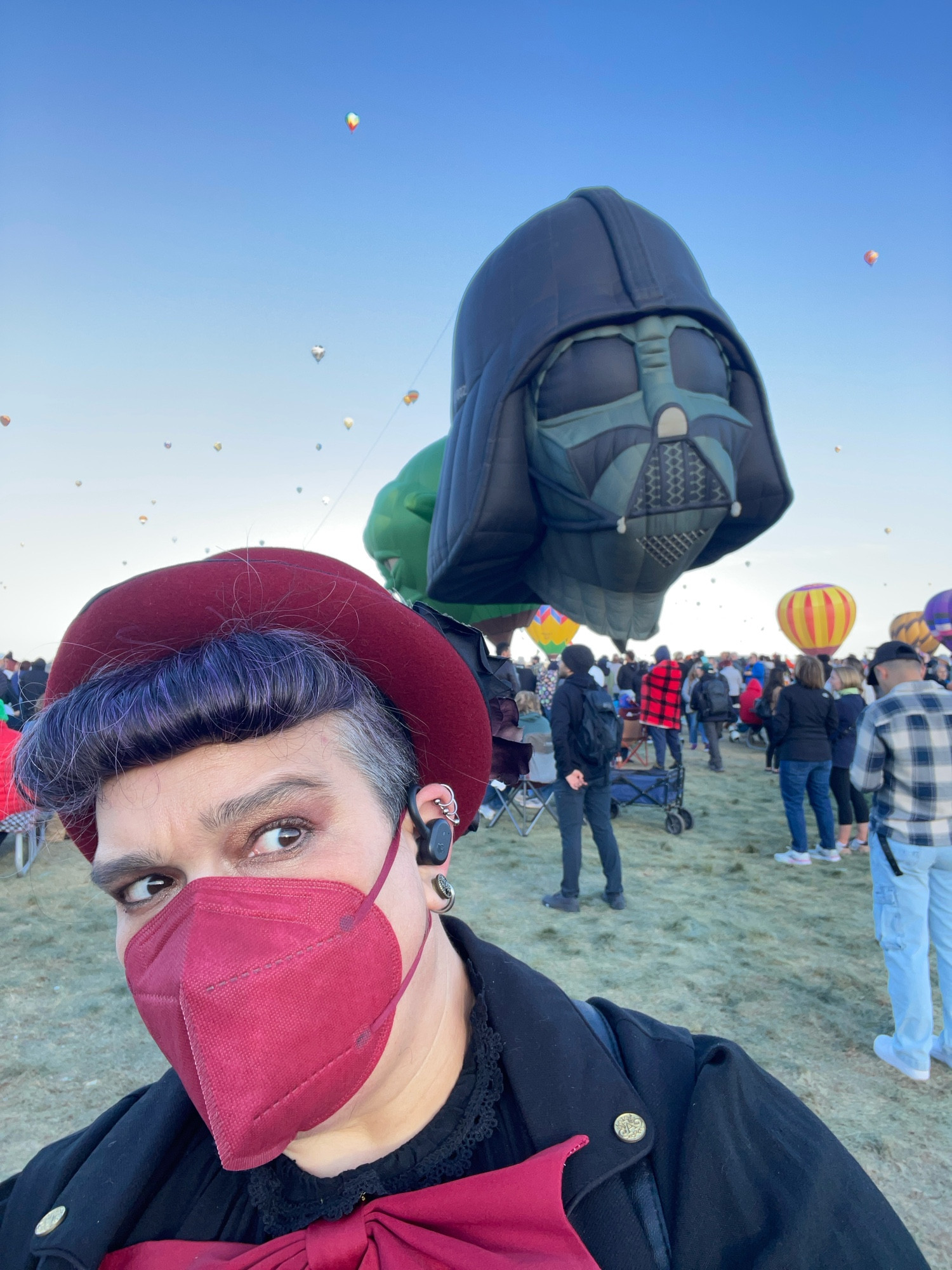 A selfie of Elya with their hair styled in bumper bangs, wearing a dark red hat and face mask, side-eyeing the Darth Vader balloon looming menacingly over their shoulder.