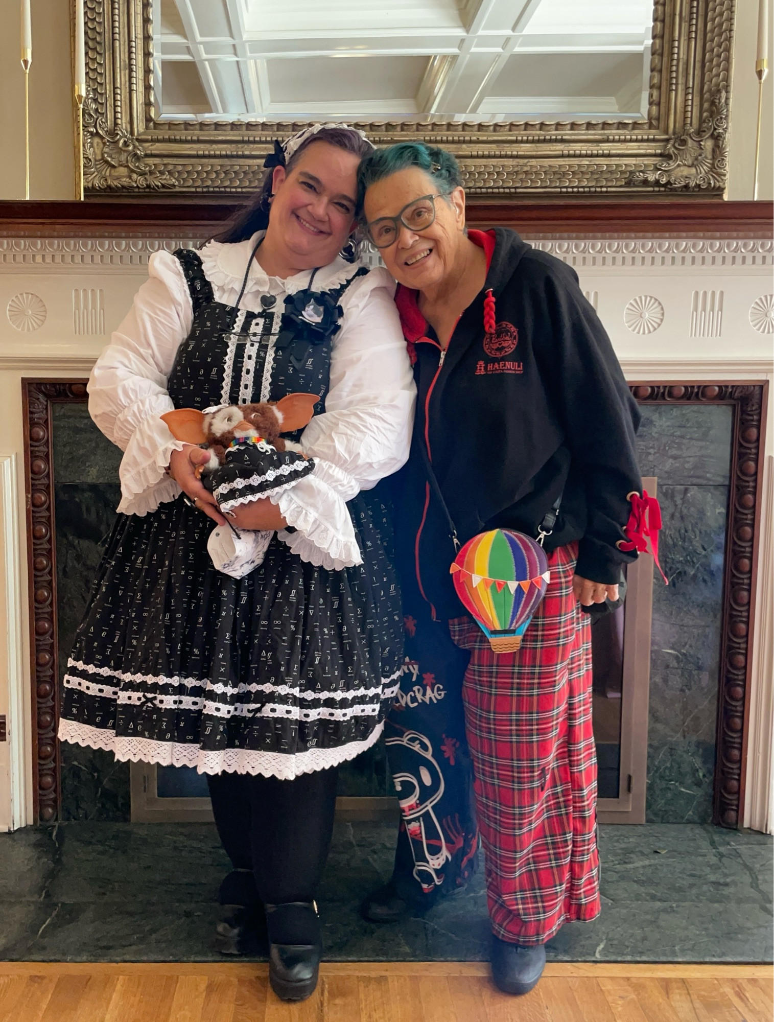 Elya and their Mom (and Gizmo) posing together in front of a marble fireplace. Elya has long purple hair and is wearing a black and white old school lolita dress printed with tiny math symbols, a white blouse with a wide "peter pan" collar and ruffled cuffs, and a black and white lacy headdress. Mom has short teal hair and is wearing a black hoodie with red ribbons laced on the sleeves, wide-leg pants where one leg is a red plaid and the other is printed with a graffiti-style illustration of Gloomy Bear, and a colorful hot-air-balloon shaped crossbody bag. Tiny fluffy mogwai Gizmo is also wearing a black and white dress, twinning with Elya.