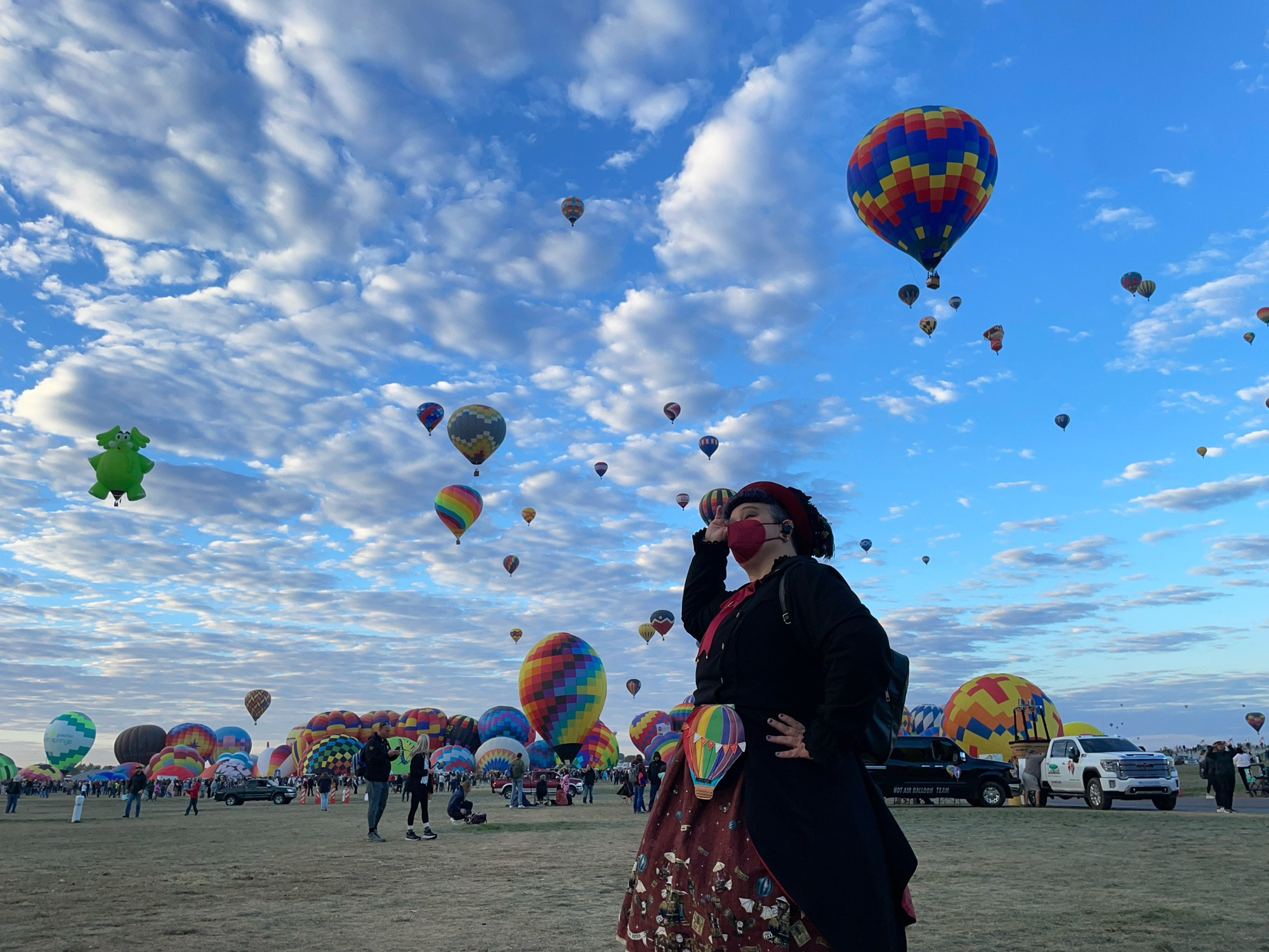 Elya looks up at a blue Albuquerque sky filled with small puffy clouds and a generous sprinkling of hot air balloons; more colorful balloons in various stages of inflation line the horizon behind them. They are wearing a steampunk style classic lolita outfit in black and dark red, with a rainbow-colored waist pouch shaped like a hot air balloon. 