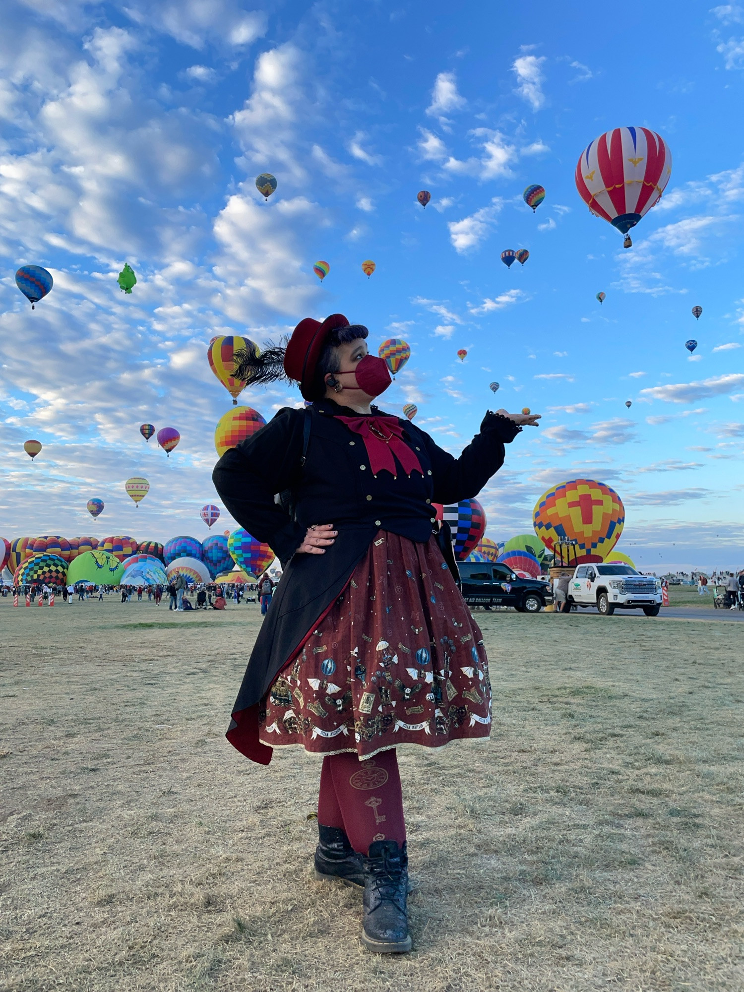 Elya poses like a teapot in front of a sky full of hot air balloons, showing off their coord. They are wearing a bell-shaped dark red skirt with a steampunk-style print (featuring more balloons/airships); black blouse and waistcoat; dark red tights, neck bow, and hat; iridescent black doc martens boots. 