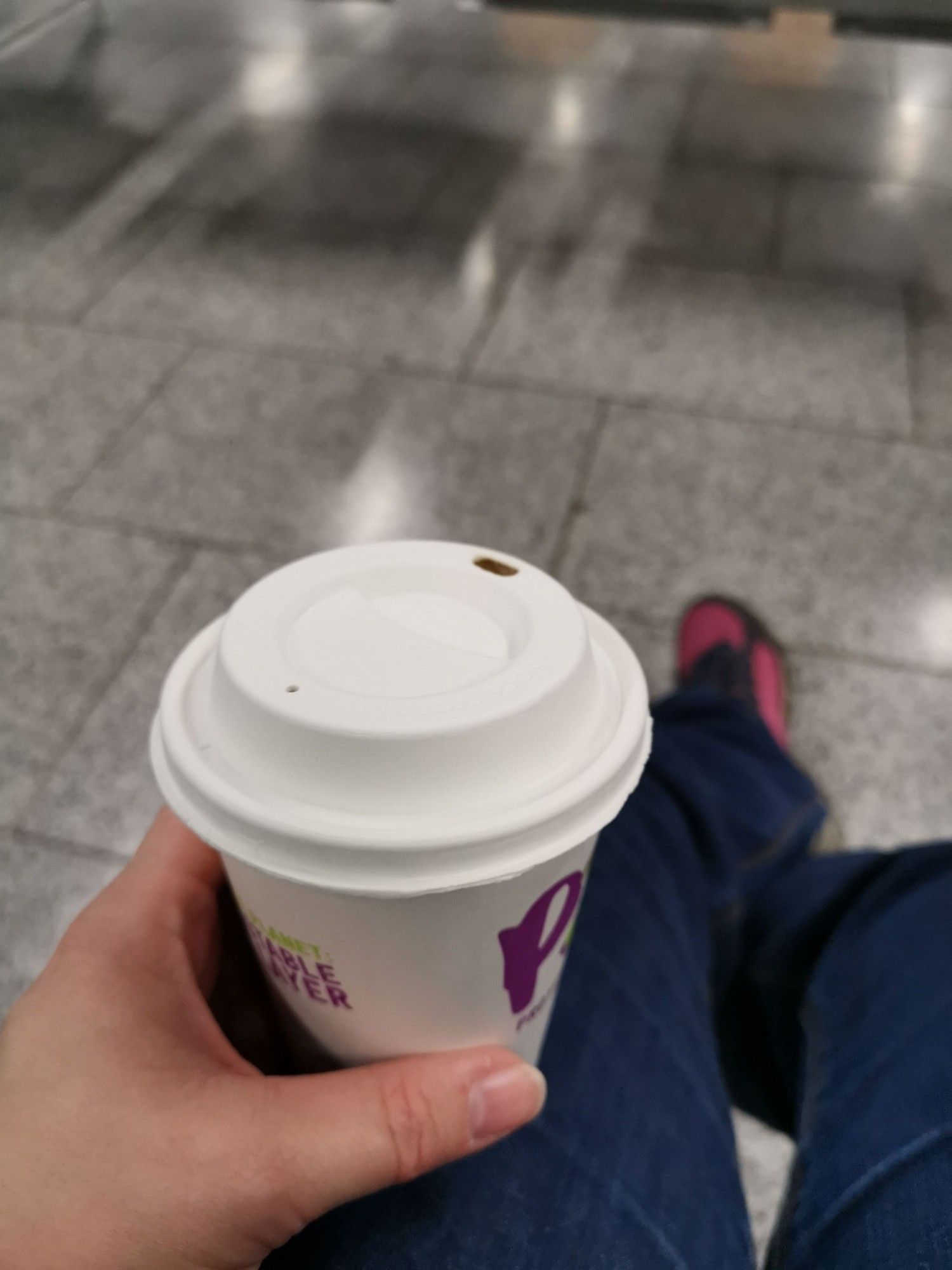 Ein Bild aus Protagonistenperspektive: ein Becher Kaffee, im Hintergrund wartende Füße.