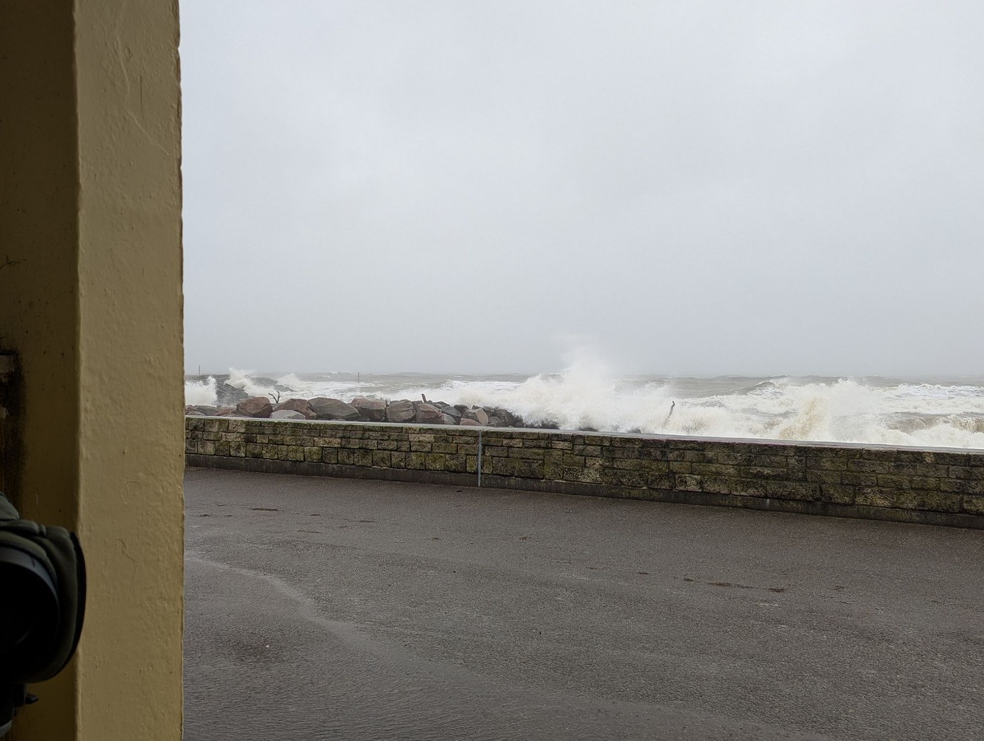 Photo taken from seafront shelter, looking over sea wall at rough sea. Objective lens of telescope just visible bottom left.