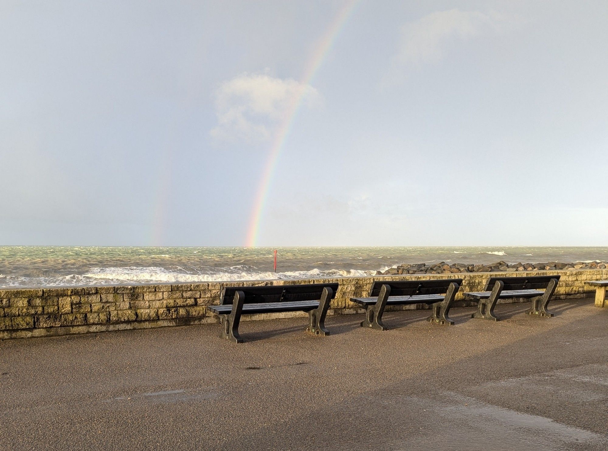 Double rainbow over the sea, viewed from seafront shelter. Benches and low seawall in foreground.