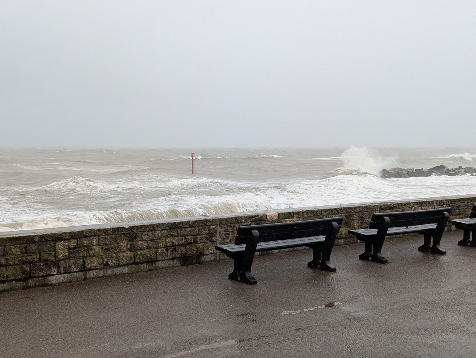 Photo of rough sea, with seawall and benches in foreground.