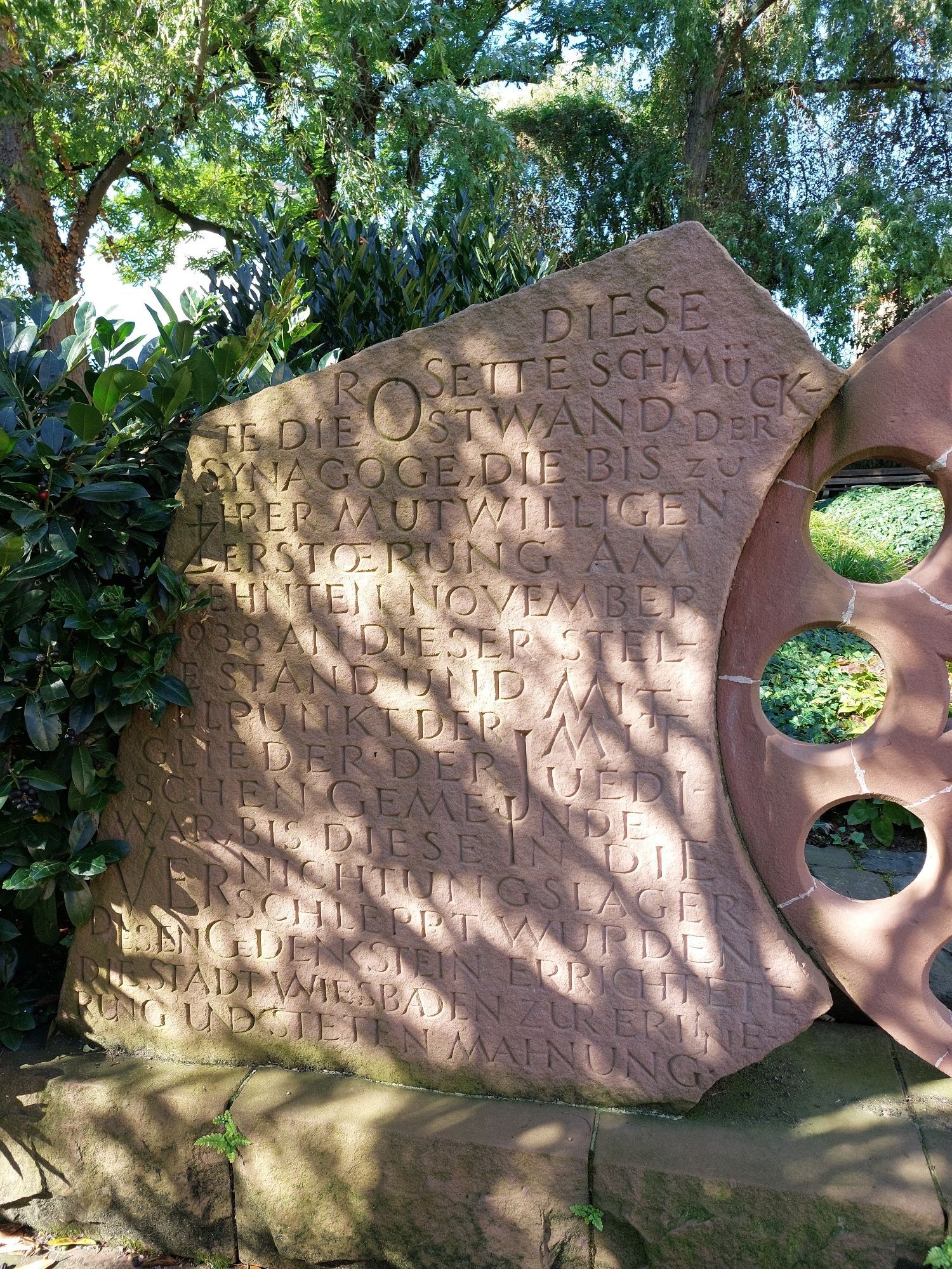 Inschrift auf der linken Seite: 
Diese Rosette schmückte die Ostwand der Synagoge, die bis zu ihrer mutwilligen Zerstörung am zehnten November 1938 an dieser Stelle stand und Mittelpunkt der Mitglieder der jüdischen Gemeinde war, bis diese in die Vernichtungslager verschleppt wurden. Diesen Gedenkstein errichtete die Stadt Wiesbaden zur Erinnerung und steten Mahnung.