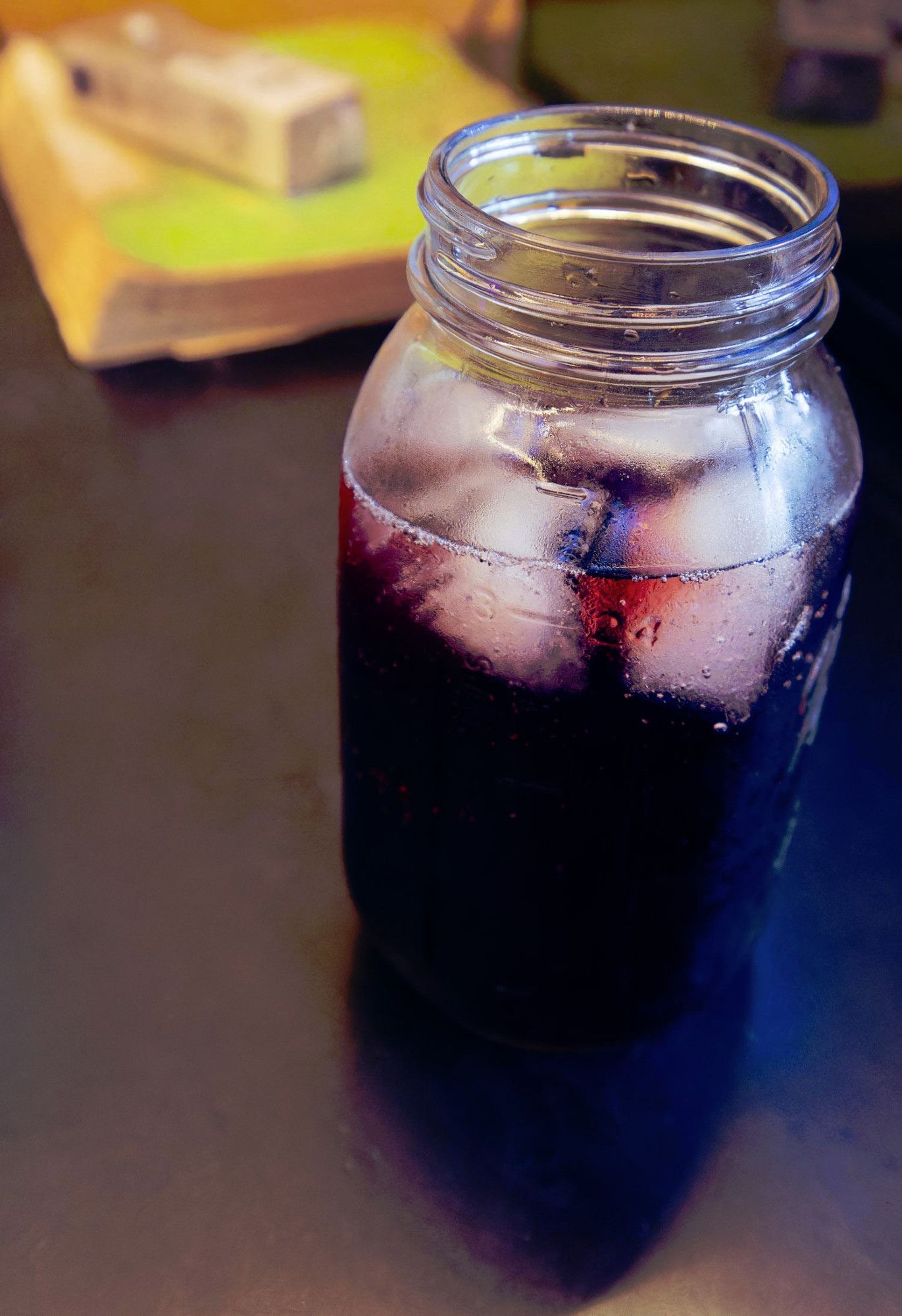 Large Mason Jar with Four Ice Cubes and a Purple Sparkly Liquid on a black tabletop with a big green softcover book and a rectangular plastic box on top of it