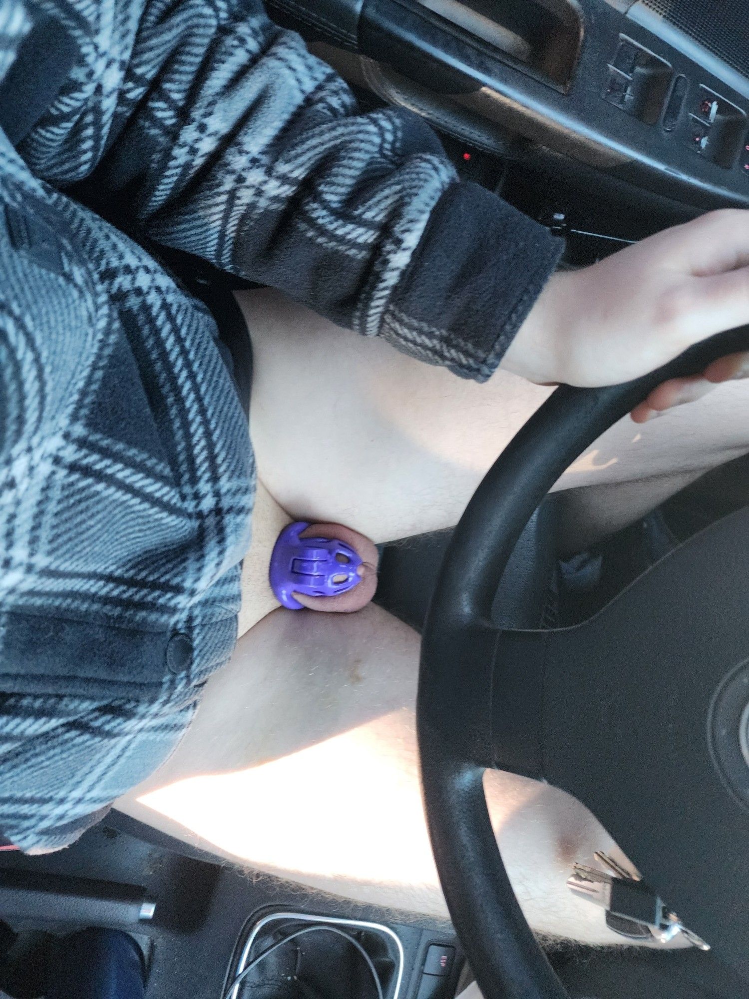 Picture of a male in a purple chastity cage sitting in the driver's seat of a car and holding the steering wheel with his left hand