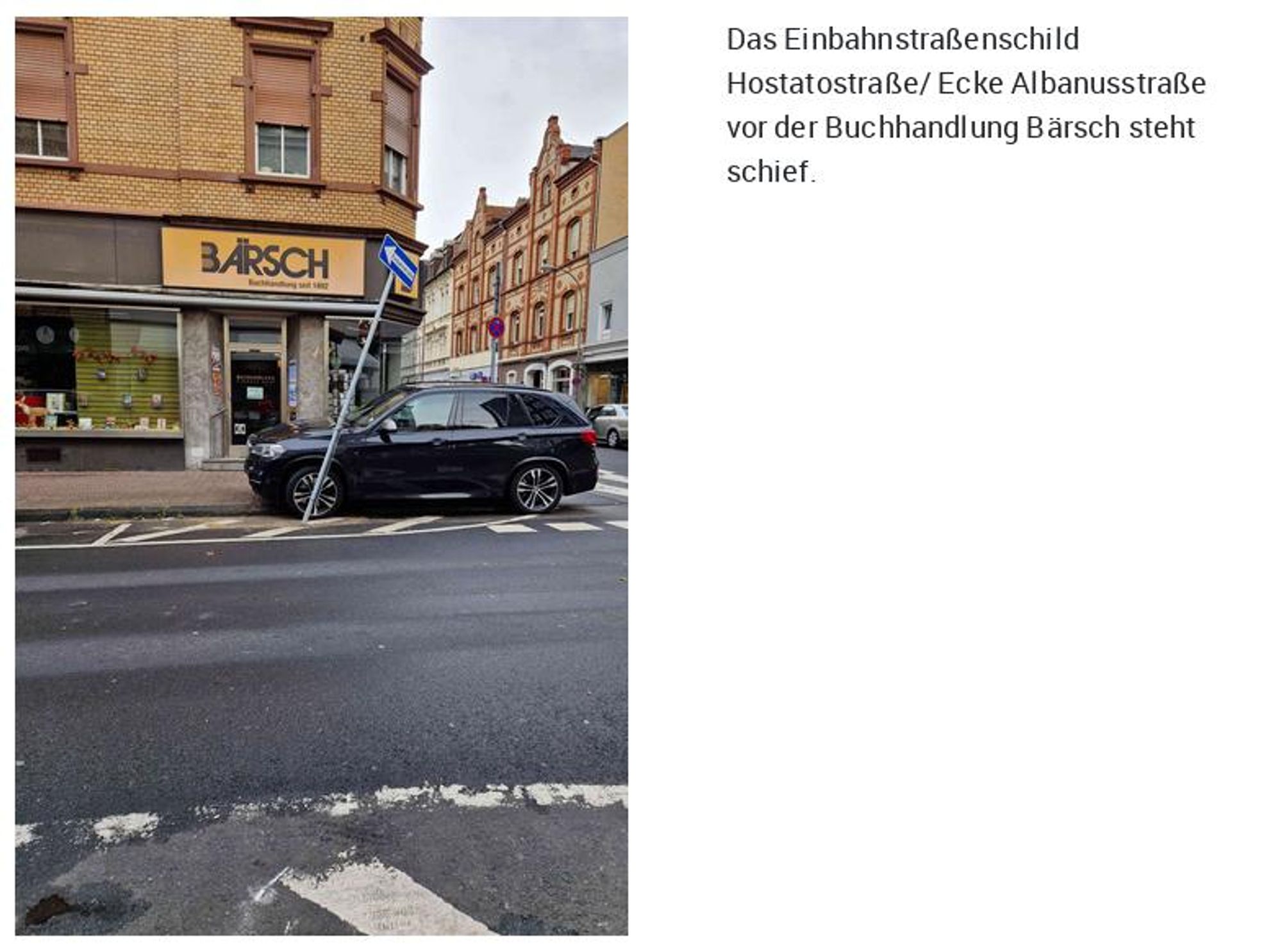 Links das Foto eines Gebäudes an einer Straßenecke. Direkt an der Ecke steht ein schwarzes Kfz und davor ein schief stehender Schildermast.
Rechts der Text "Das Einbahnstraßenschild Hostatostraße/ Ecke Albanusstraße vor der Buchhandlung Bärsch steht schief."