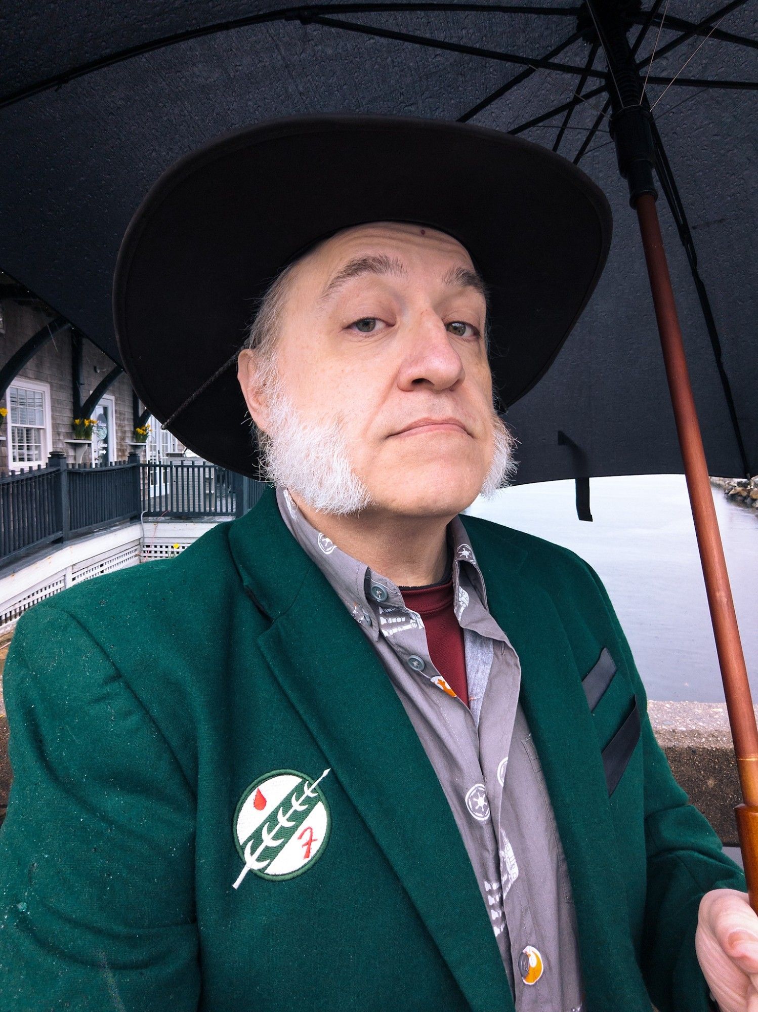 Outdoor selfie on a rainy day. I'm holding an umbrella. I'm wearing a blazer styled after Boba Fett's armor, complete with his signature sigil.