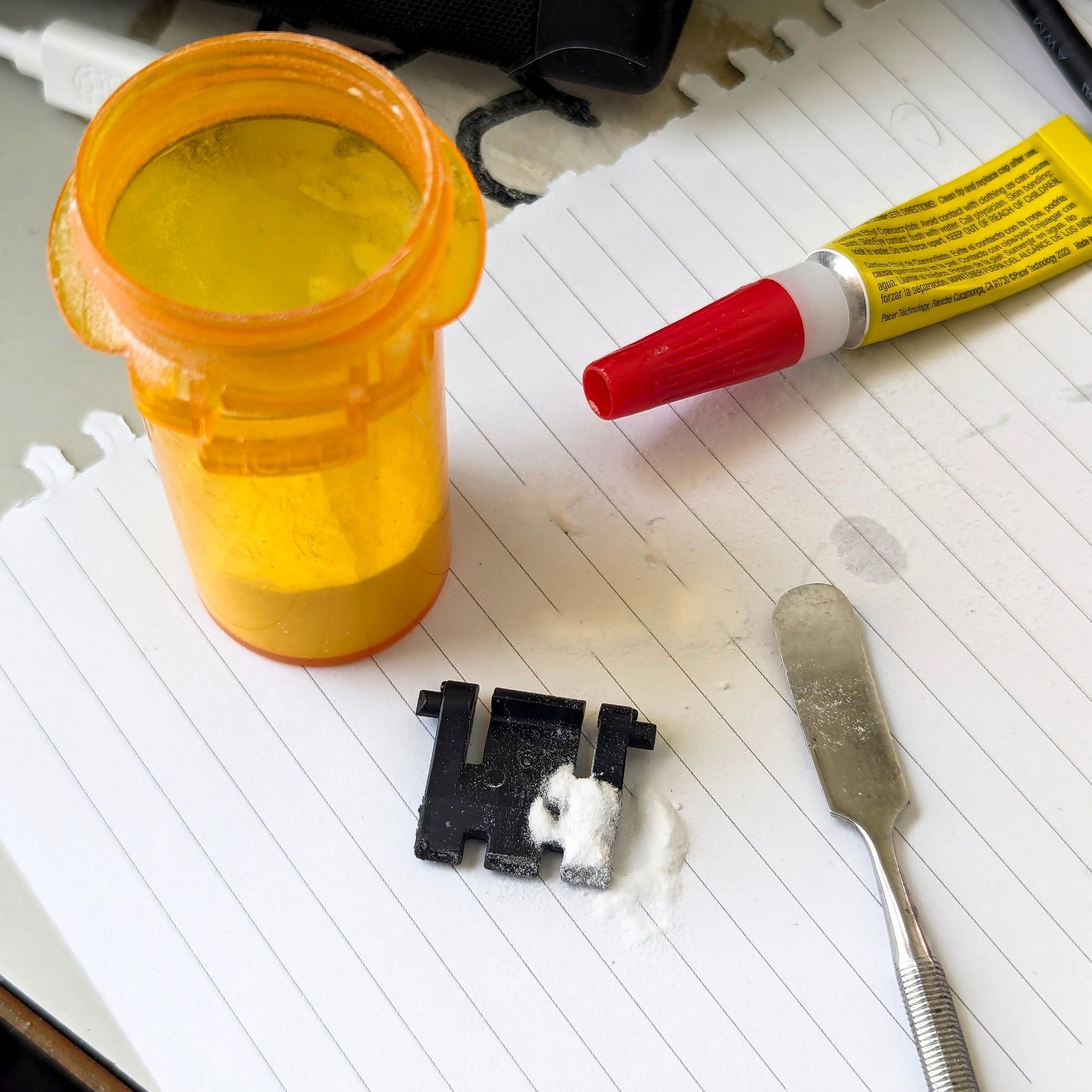 A closeup of items on a desk. At center is a black plastic part being mended. A corner of the part is covered with a small amount of white powder. Arranged around the part are a prescription pill bottle half-filled with white powder, a tube of cyanoacrylate glue, and a dental tool resembling a small spatula.