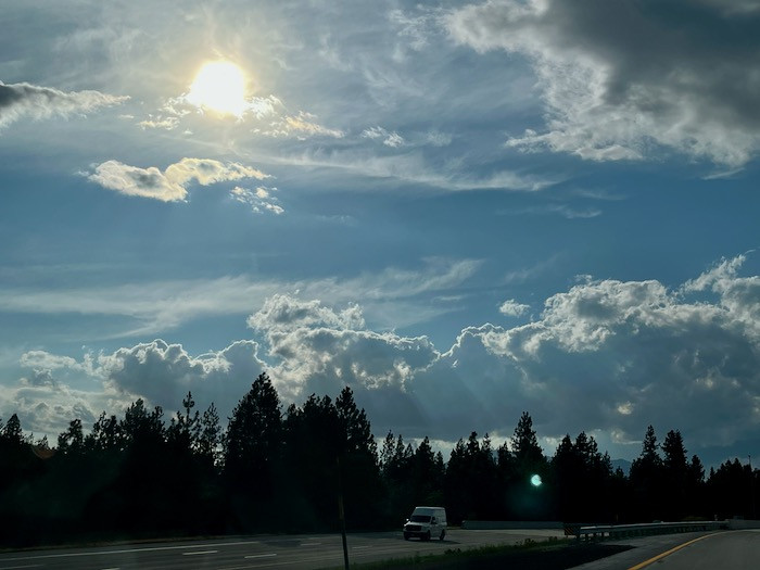 photo: the sun in the west shining through layers of wispy clouds, above other layers of chunky cloud in a grey-blue sky. Taken from highway 5 with a line of dark evergreens on the side of the road.
