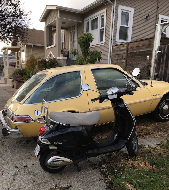photo: pale yellow AMC Pacer parked in a residential driveway next to a a navy blue Vespa LX150.