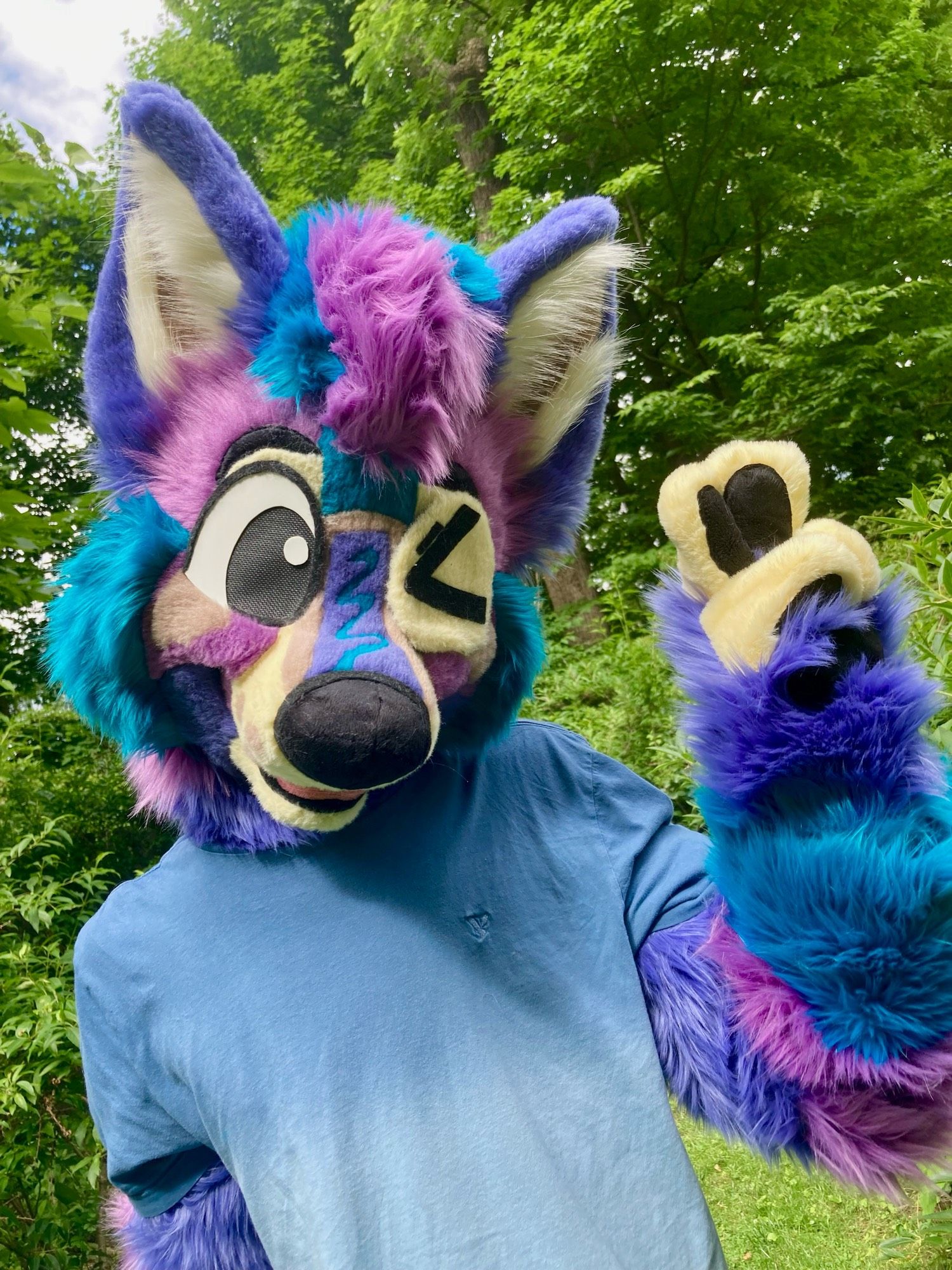 Photo of a happy toony poptart inspired wolf partial fursuit. He is winking and holding up a peace sign. The background is outdoors with lots of sunlight, lush plants, and trees.