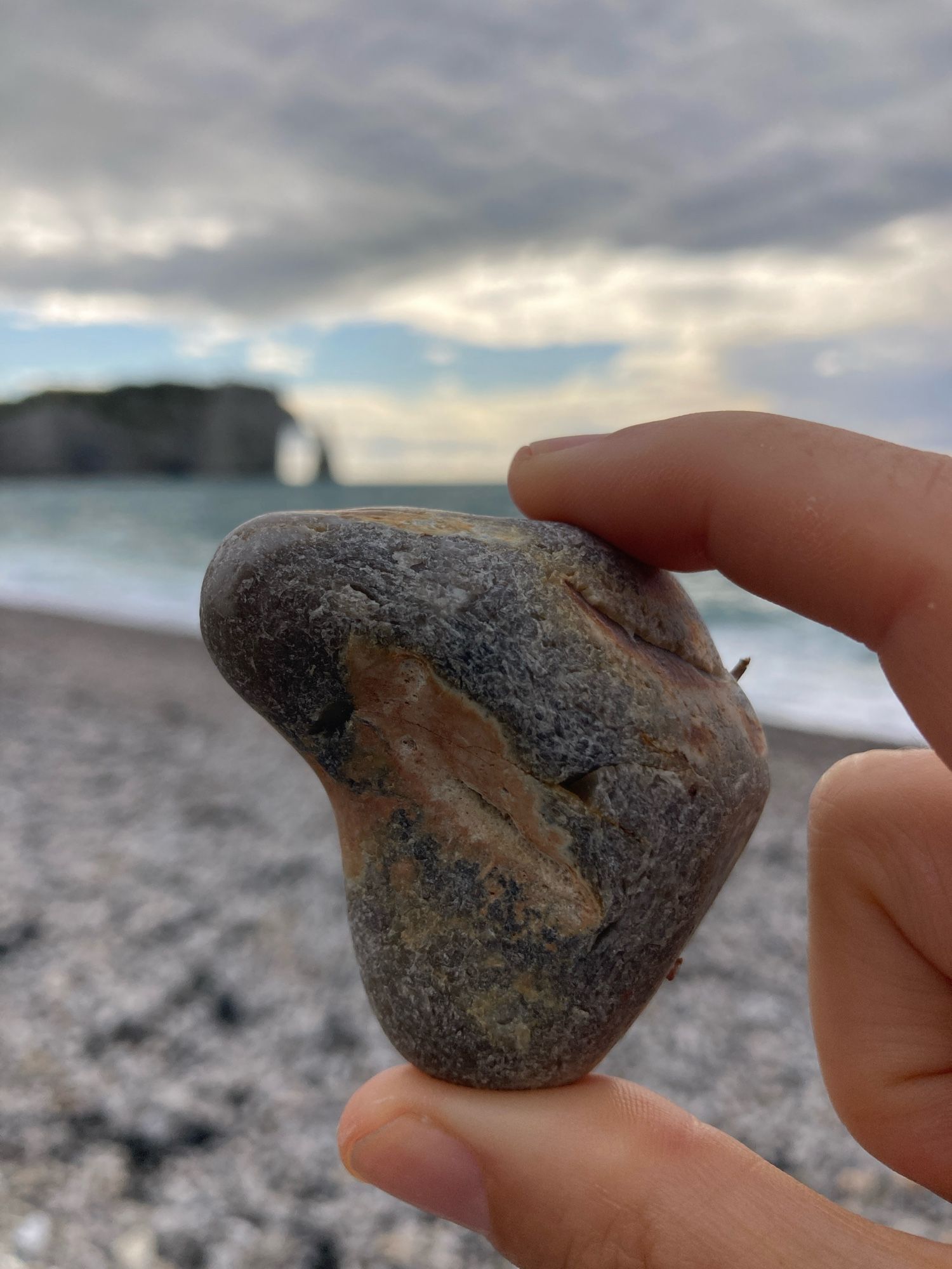 A hand holds an irregularly shaped, black and light brown beach pebble. The backdrop is a rocky beach, the sea, the sky, and the Étretat cliff and arch in the background.