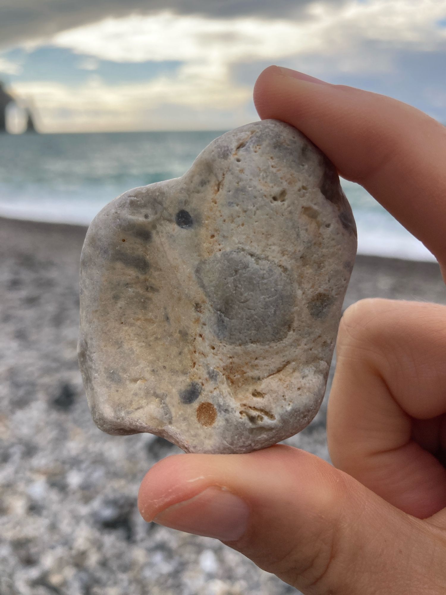 A hand holds a large, irregularly shaped, multicolored beach pebble. The backdrop is a rocky beach, the sea, the sky.