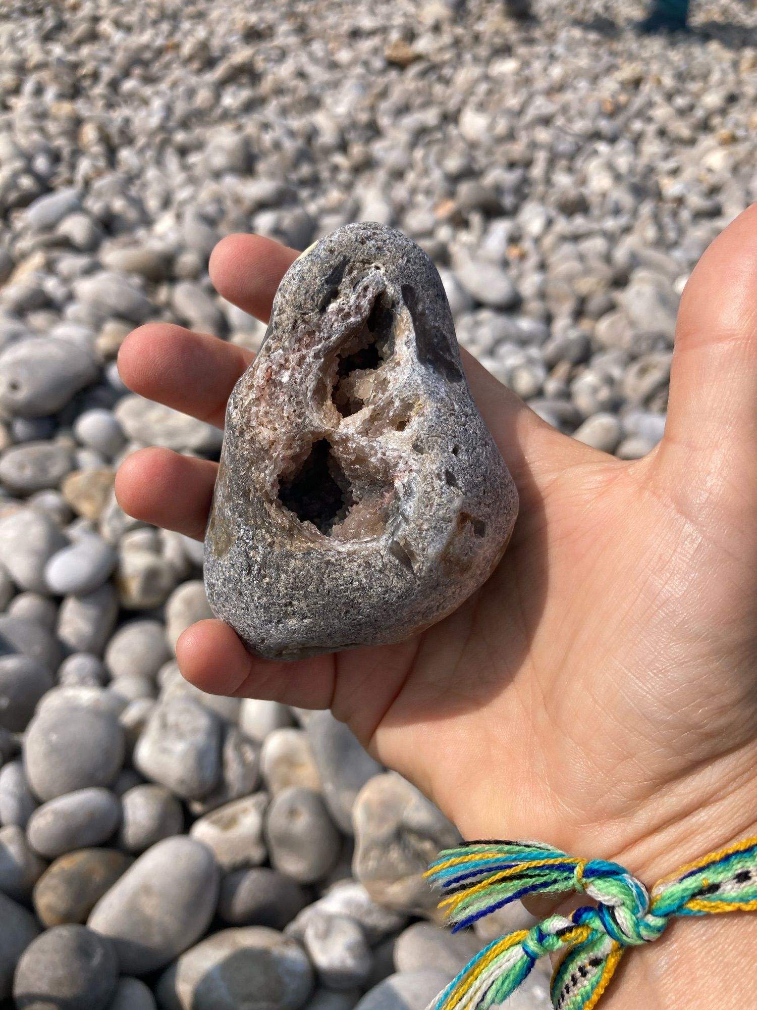 A hand holds a b shaped, brownish grey beach pebble with large connected holes in the middle in which light brown crystals glimmer. The backdrop is a rocky beach.