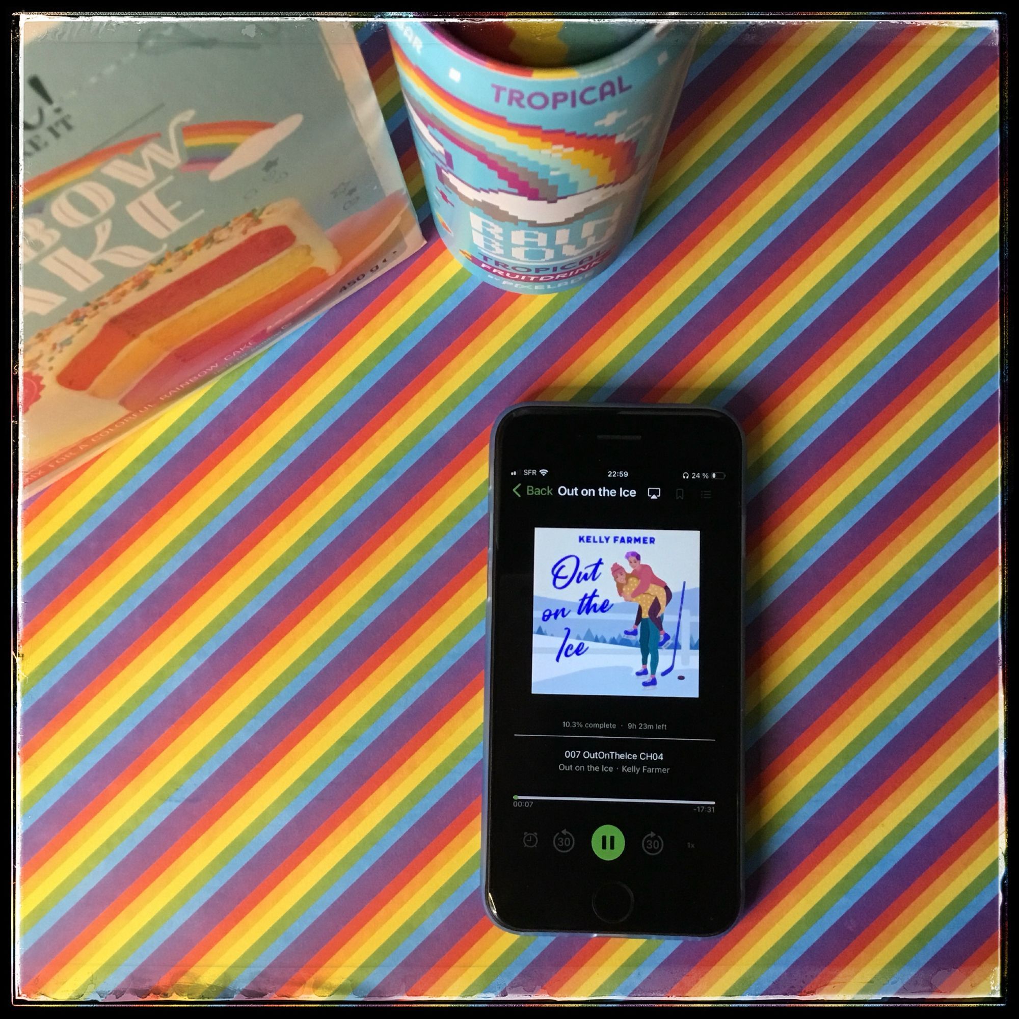 On a rainbow striped background, an iPhone with the cover of Out on the Ice by Kelly Farmer, narrated by Chelsea Stephens, with partial views of a box of rainbow cake mix and a can of rainbow tropical fruit drink.