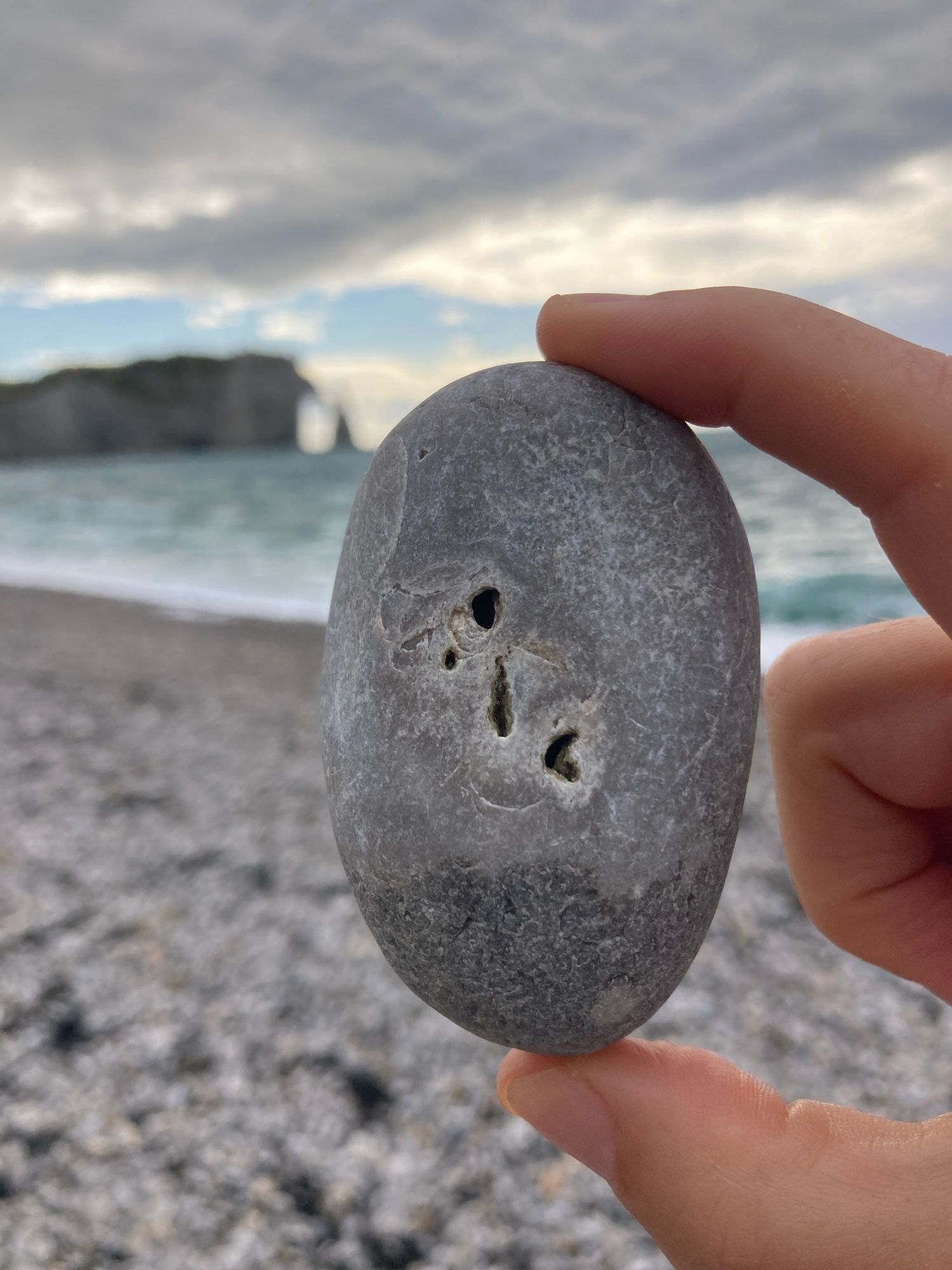 A hand holds an oval grey beach pebble with four holes towards the center, in different sizes and shapes. The backdrop is a rocky beach, the sea, the sky, and the Étretat cliff and arch in the background.