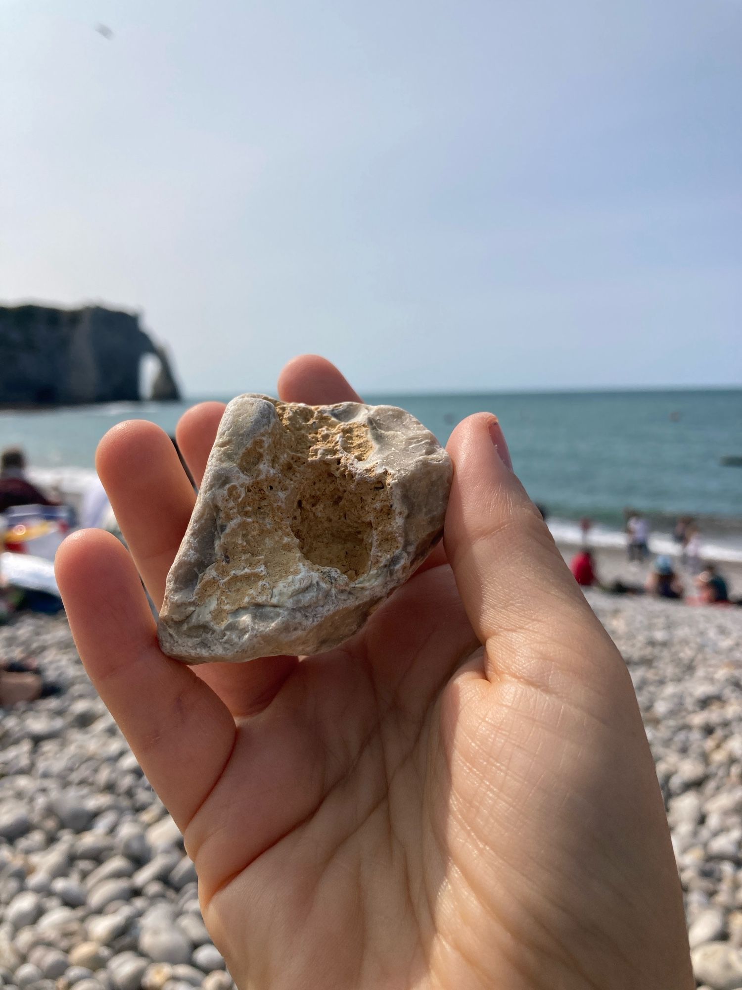 A hand holds an irregularly shaped, multicolored, beach pebble, with one straight side. The main colors are yellow, grey, white. The backdrop is a busy rocky beach, with the Étretat cliff and arch in the background.