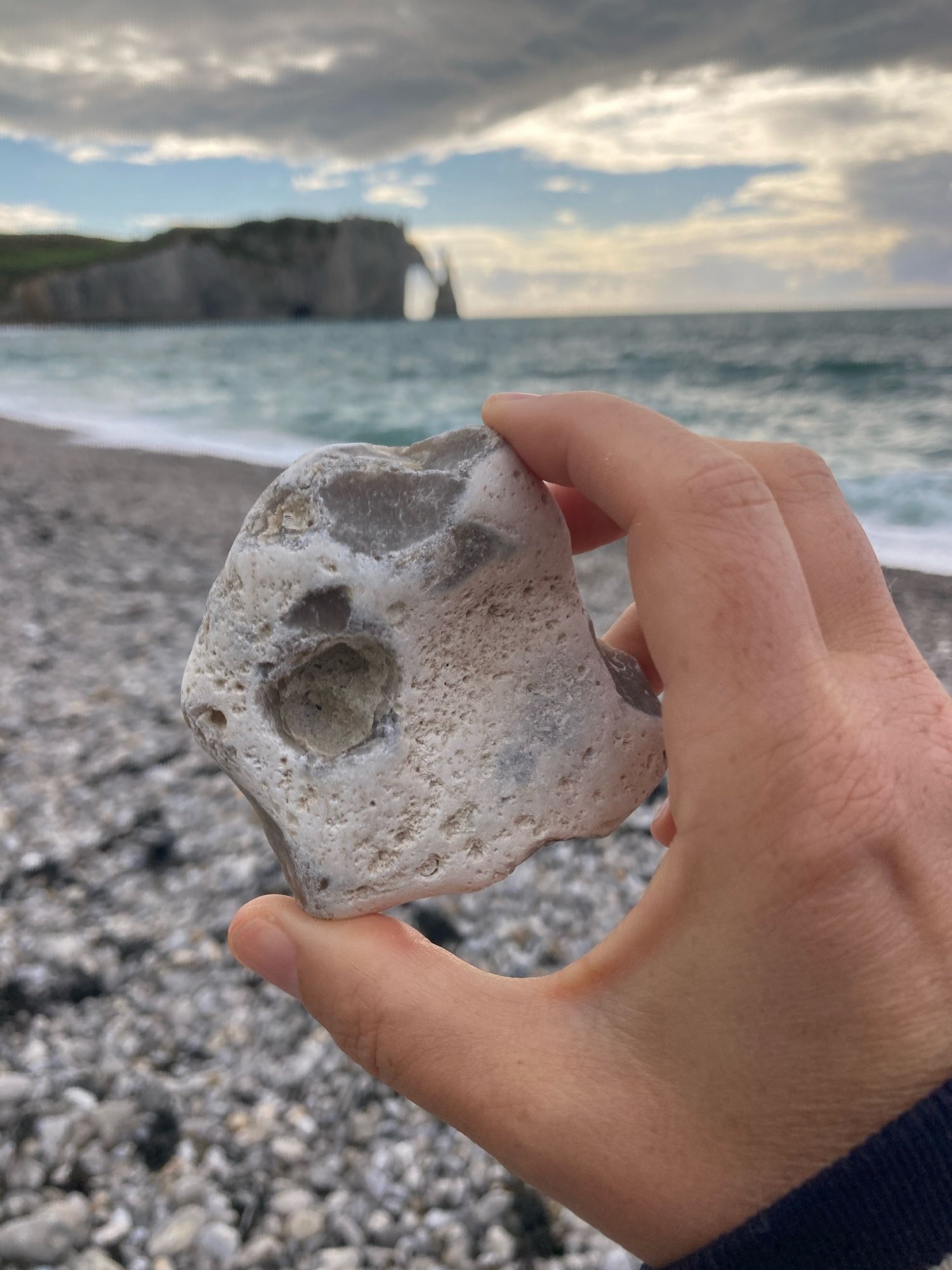 A hand holds an irregularly shaped, white beach pebble, chipped in places showing grey. The backdrop is a rocky beach, the sea, the sky, and the Étretat cliff and arch in the background.