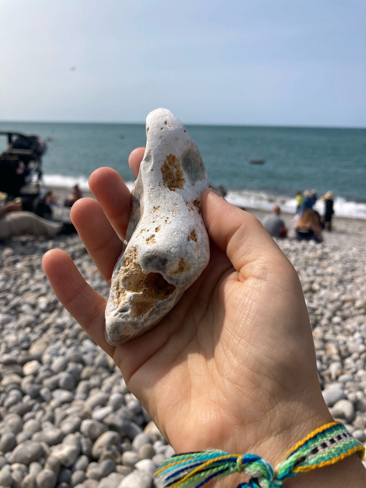 A hand holds a lost, irregularly shaped, mostly white beach pebble, with orange areas and holes. The backdrop is a busy rocky beach.